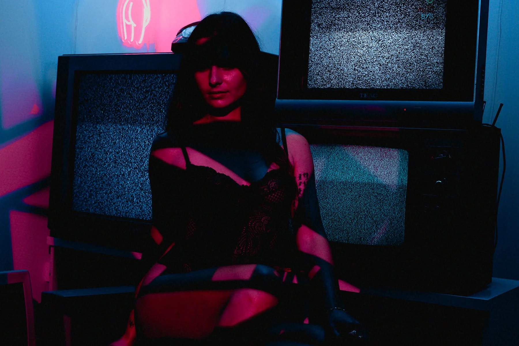 A woman in dark clothing with tattoos and fishnet gloves sitting in front of multiple television screens displaying static. The scene is illuminated with pink and blue neon lighting.