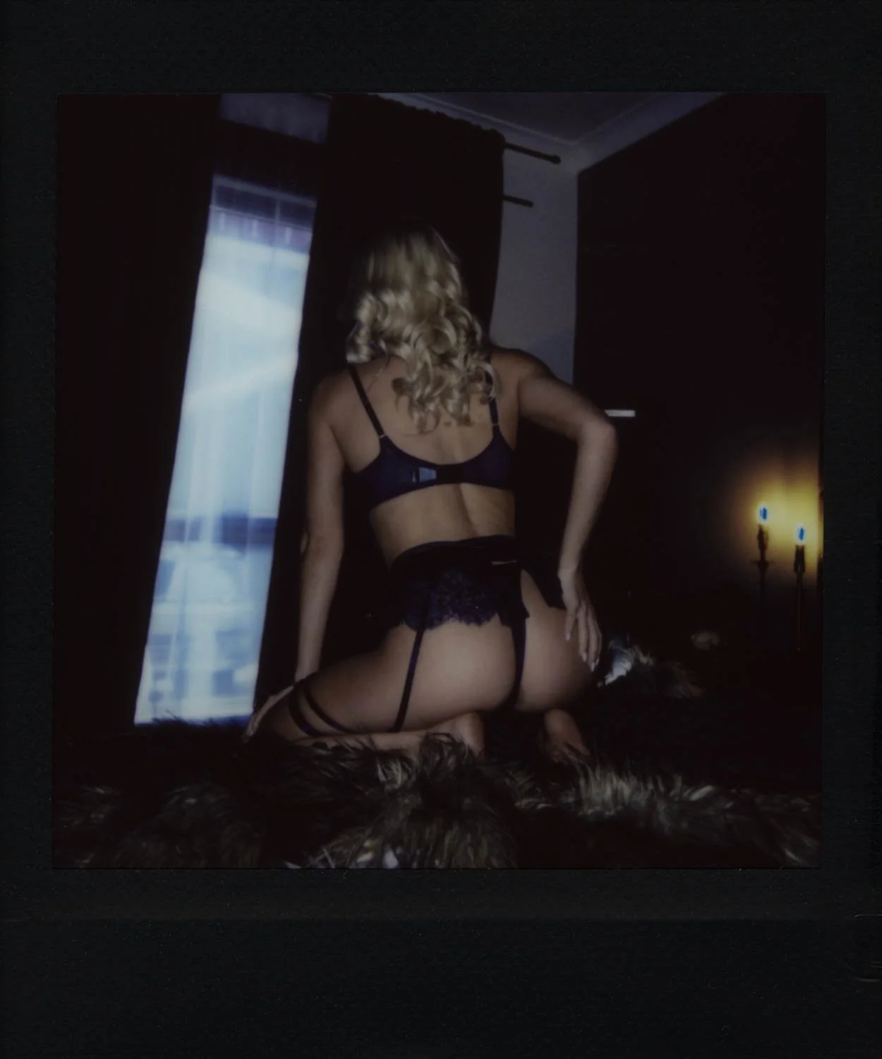 Back view instant film of model kneeling on a fur throw, wearing matching blue lingerie and garter belt. Candlelit ambience and dark drapery produce decadent, cinematic mood.