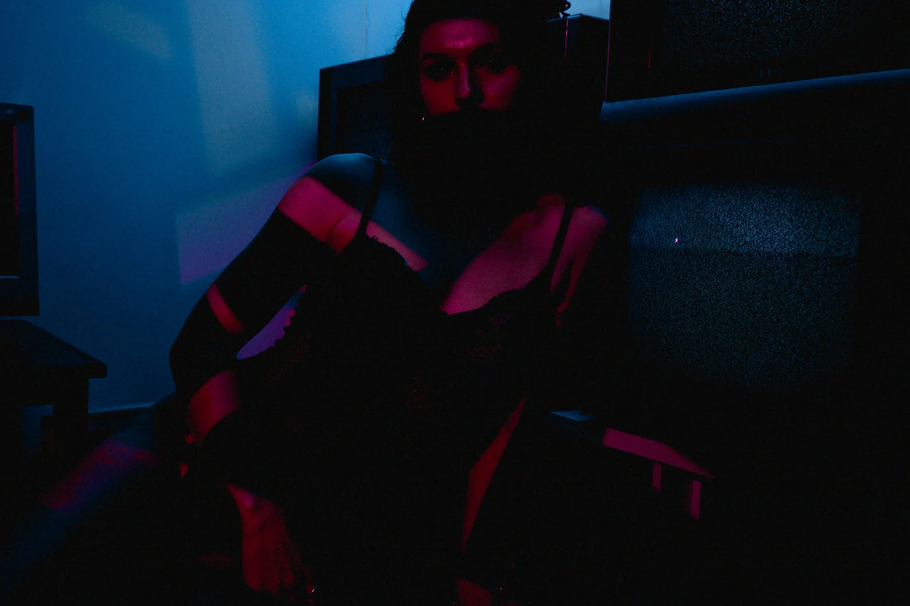 A woman with dark hair and makeup wearing a black and pink striped outfit in a dark room illuminated by blue and pink neon lights.