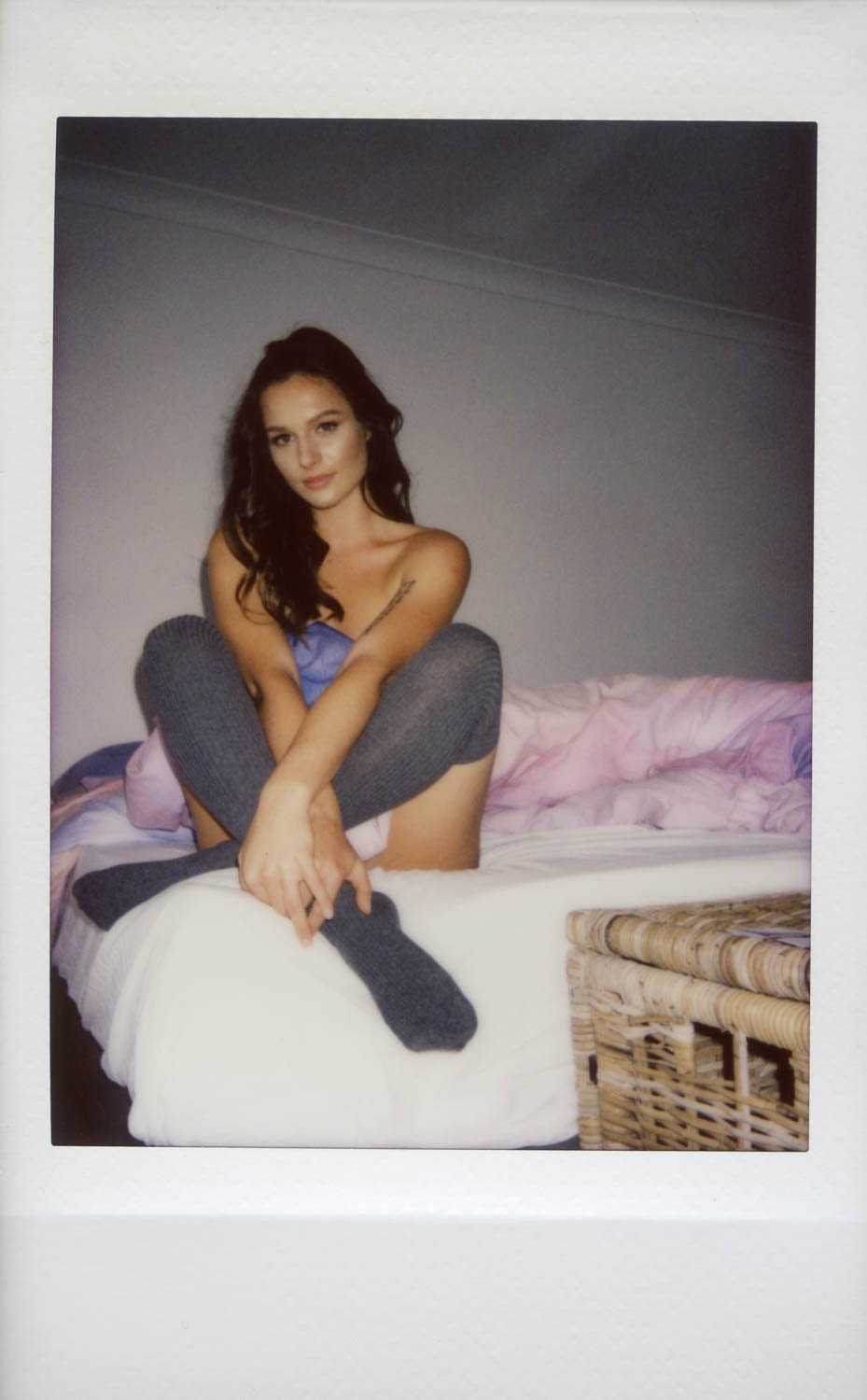 Young woman sitting on bed with crossed legs, wearing a blue top and gray socks, in a bedroom with a pink and purple blanket and wicker furniture.