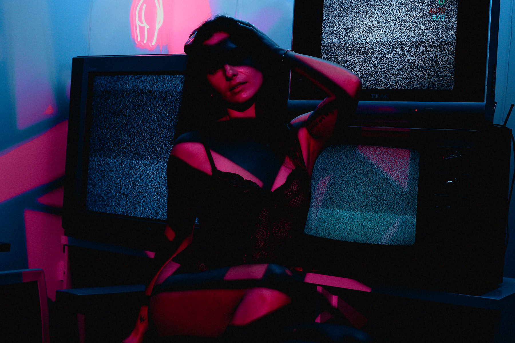 A woman with dark hair wearing black lingerie, sitting in front of two CRT televisions displaying static. The scene is illuminated with pink and blue neon lighting, creating a cyberpunk aesthetic.