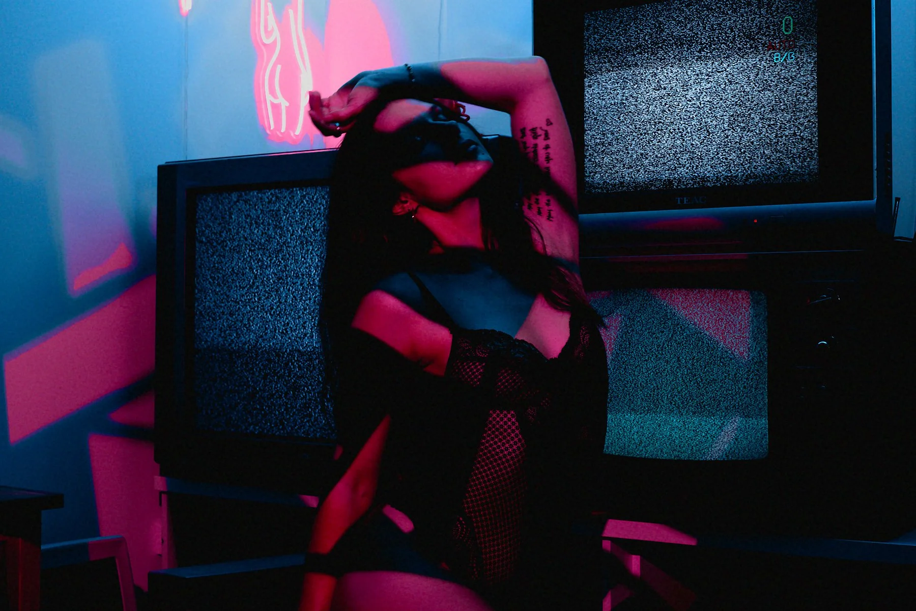 A woman wearing black lingerie, fishnet gloves, and a bra sitting in front of two old television screens displaying static noise, with pink and blue neon lighting creating an atmosphere fitting a retro or cyberpunk aesthetic.