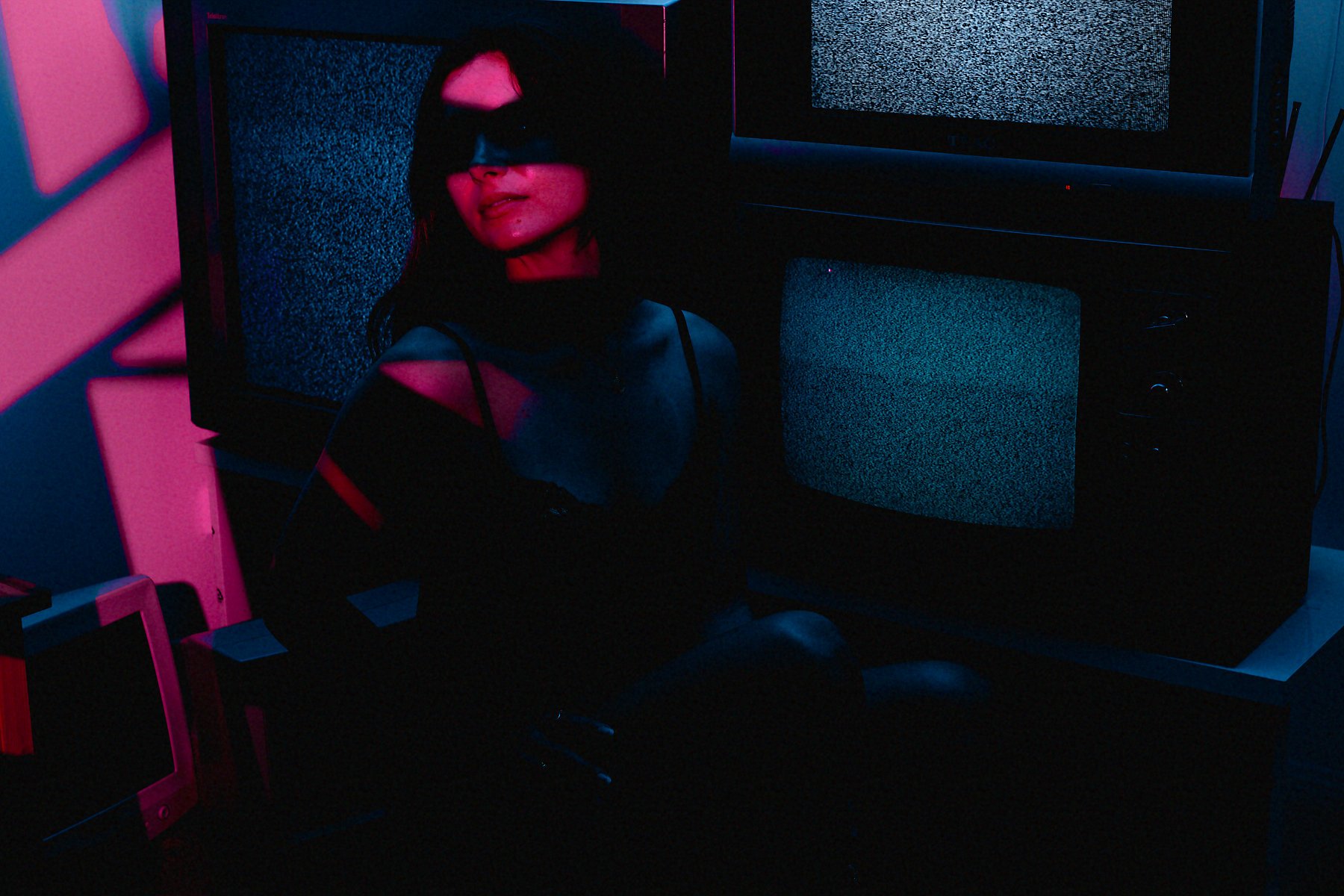 A woman with dark hair wearing sunglasses and a black top with red straps, sitting in a dark room with pink and blue neon lighting. Behind her, there are two vintage television sets with static screens, and there is a pink chair in the foreground.