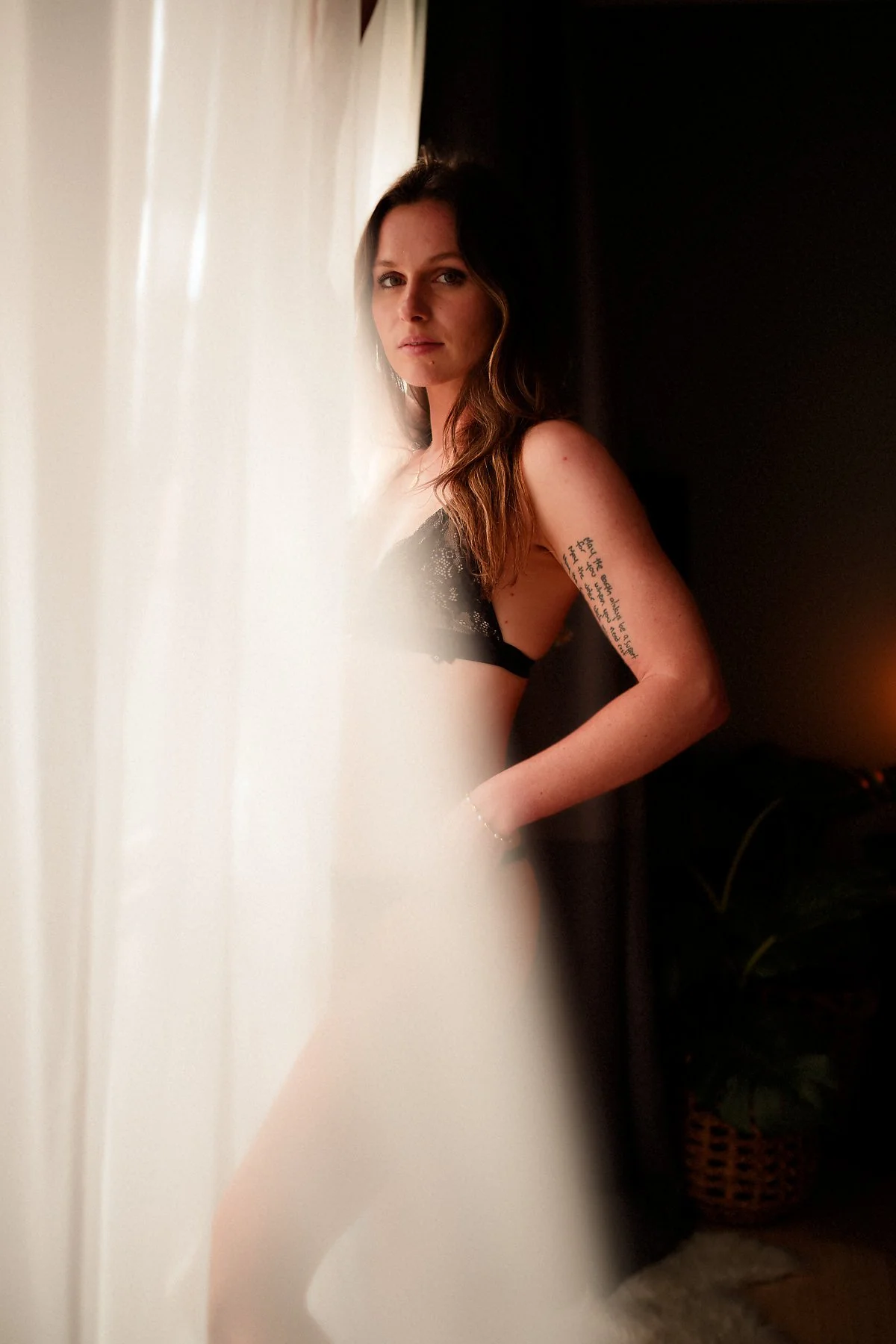 A woman with long, wavy brown hair leaning against a wall, wearing a black lace top, with a tattoo on her right arm and a neutral facial expression.