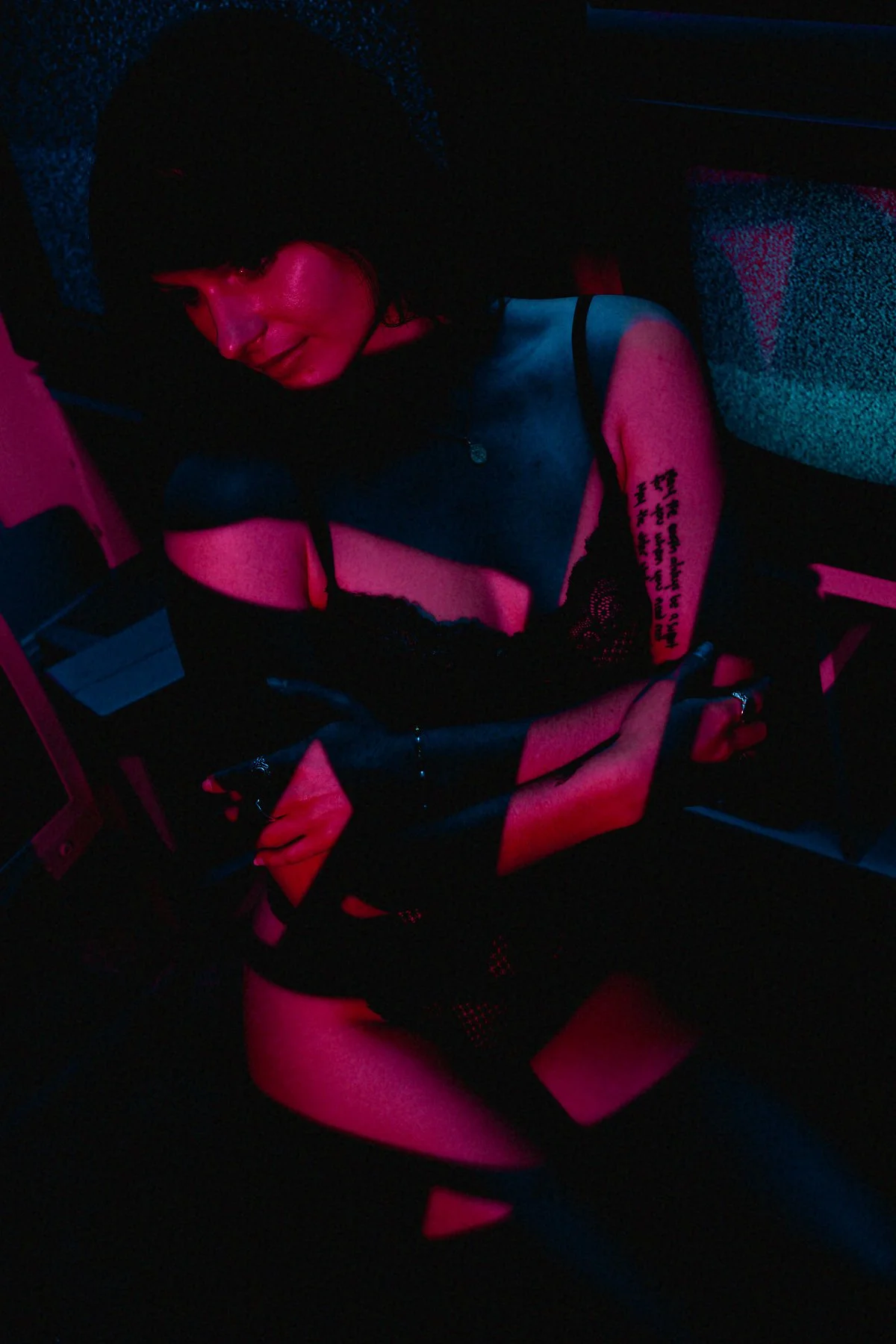 A person sitting in a dark room illuminated with pink and blue light, wearing a black tank top with tattoos on the arm, a ring, and a necklace, with their head tilted downward.
