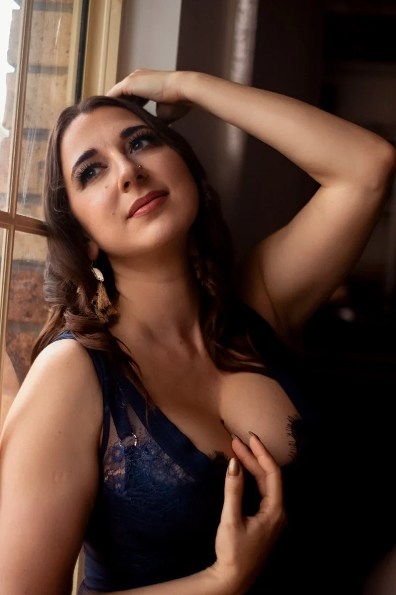 A woman with long brown hair, wearing a dark blue dress, posing with one arm raised behind her head and her hand resting on her forehead, looking thoughtfully out a window.