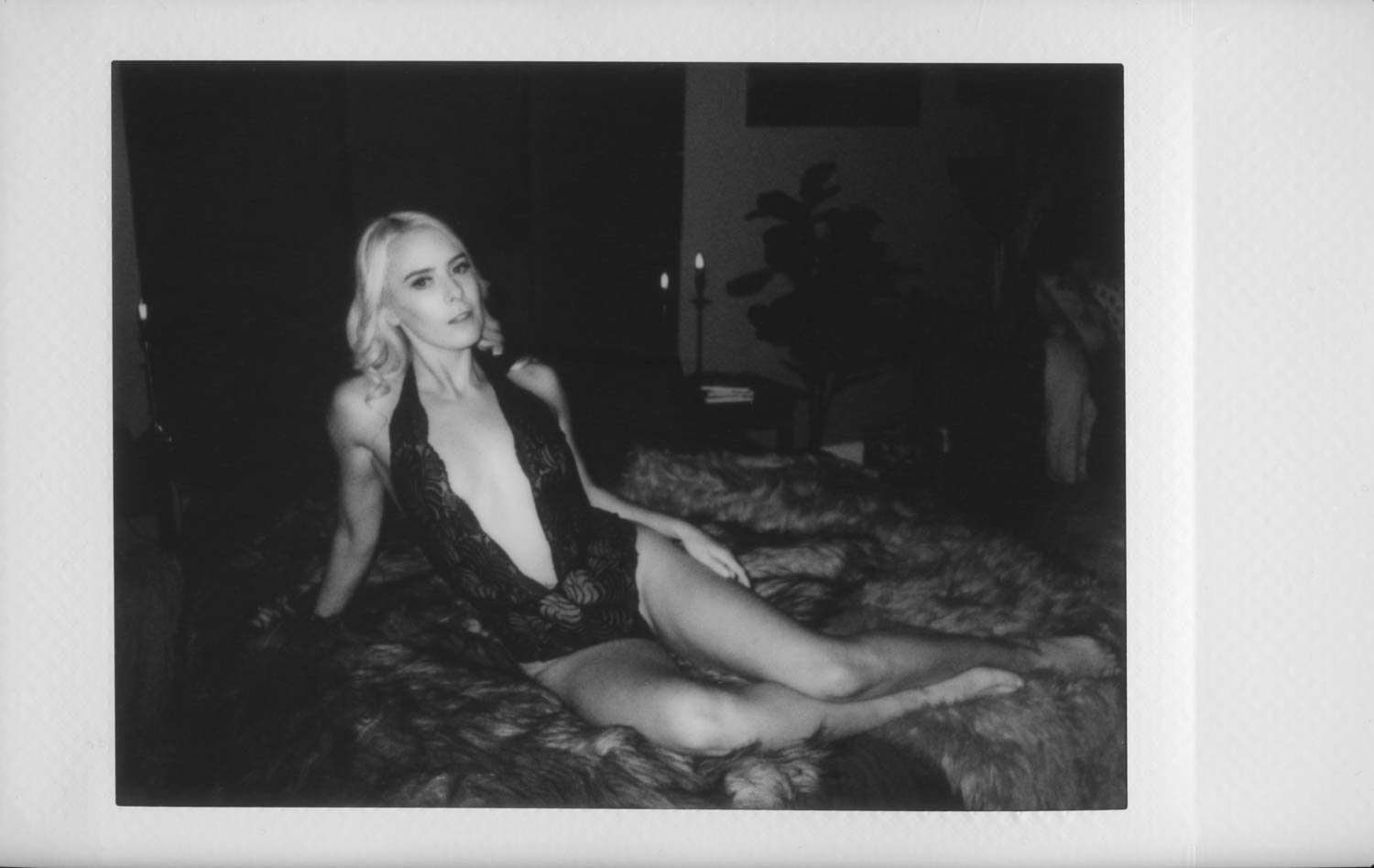 Black and white photo of a woman with blonde hair in lingerie, sitting on a bed in a dimly lit room.