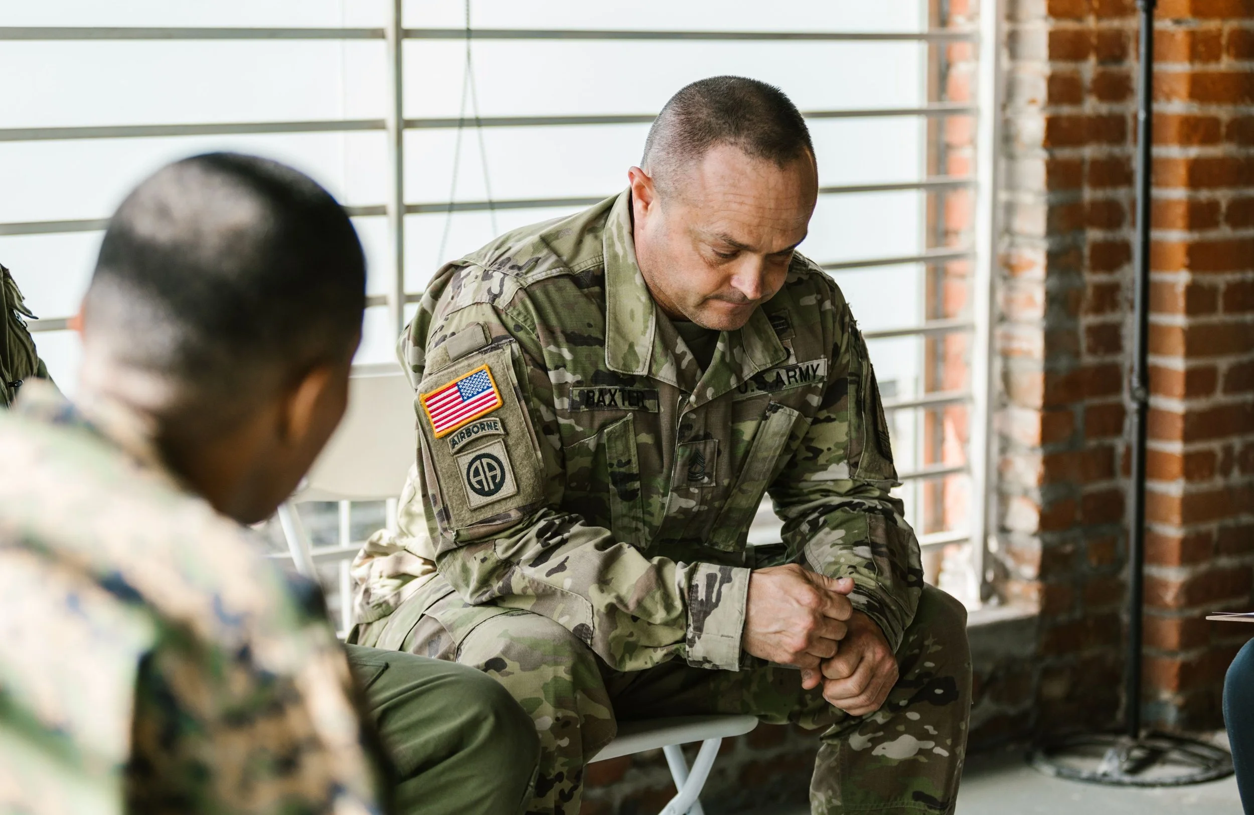 Military officer in camouflage uniform sitting with head bowed, hands clasped, in an indoor space with brick walls and metal railing window.