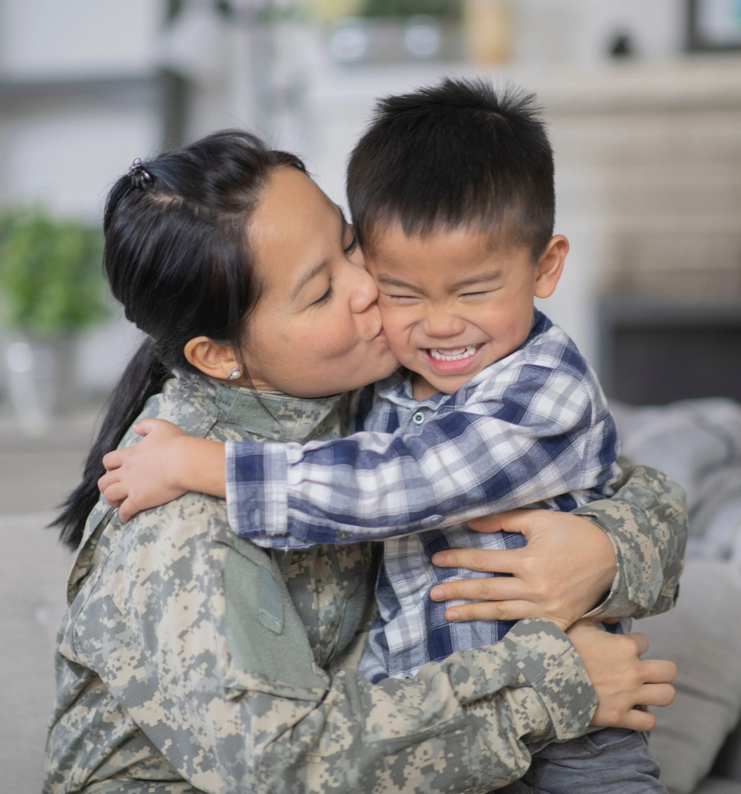 A woman in military uniform embracing and kissing a young boy in a plaid shirt, both smiling and showing affection.