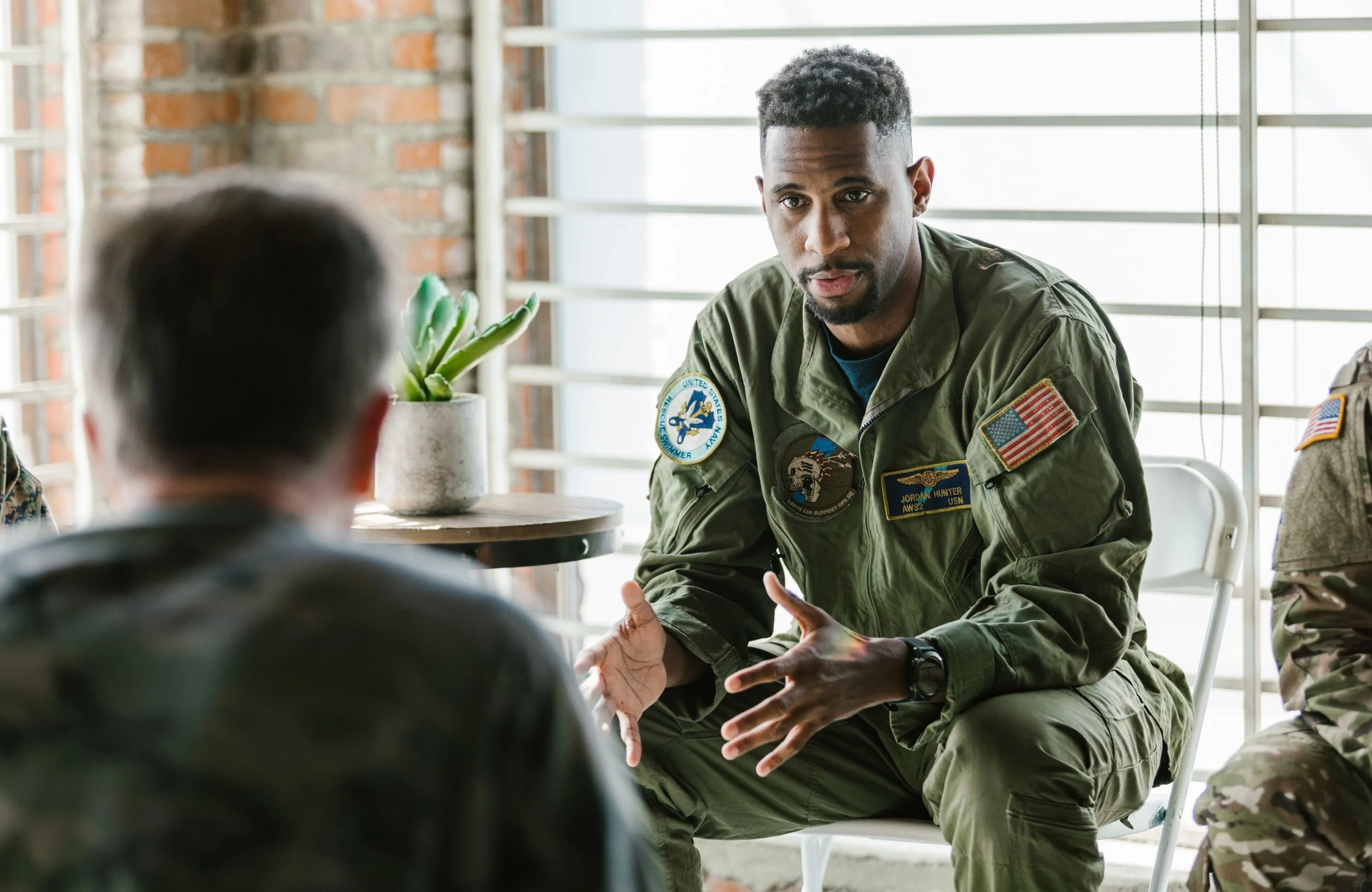 A man in military uniform engaging in a serious conversation with a person sitting across from him in a room with brick walls and large windows.