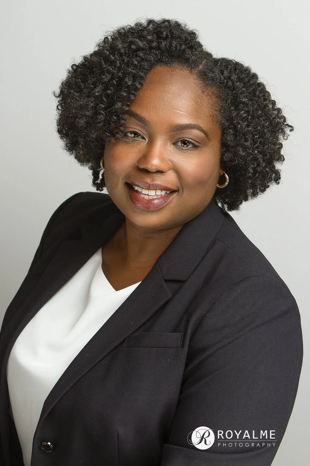 A woman with dark curly hair, wearing a black blazer and a white top, smiling, against a plain light background. Logo of Royalme Photography in the bottom right corner.