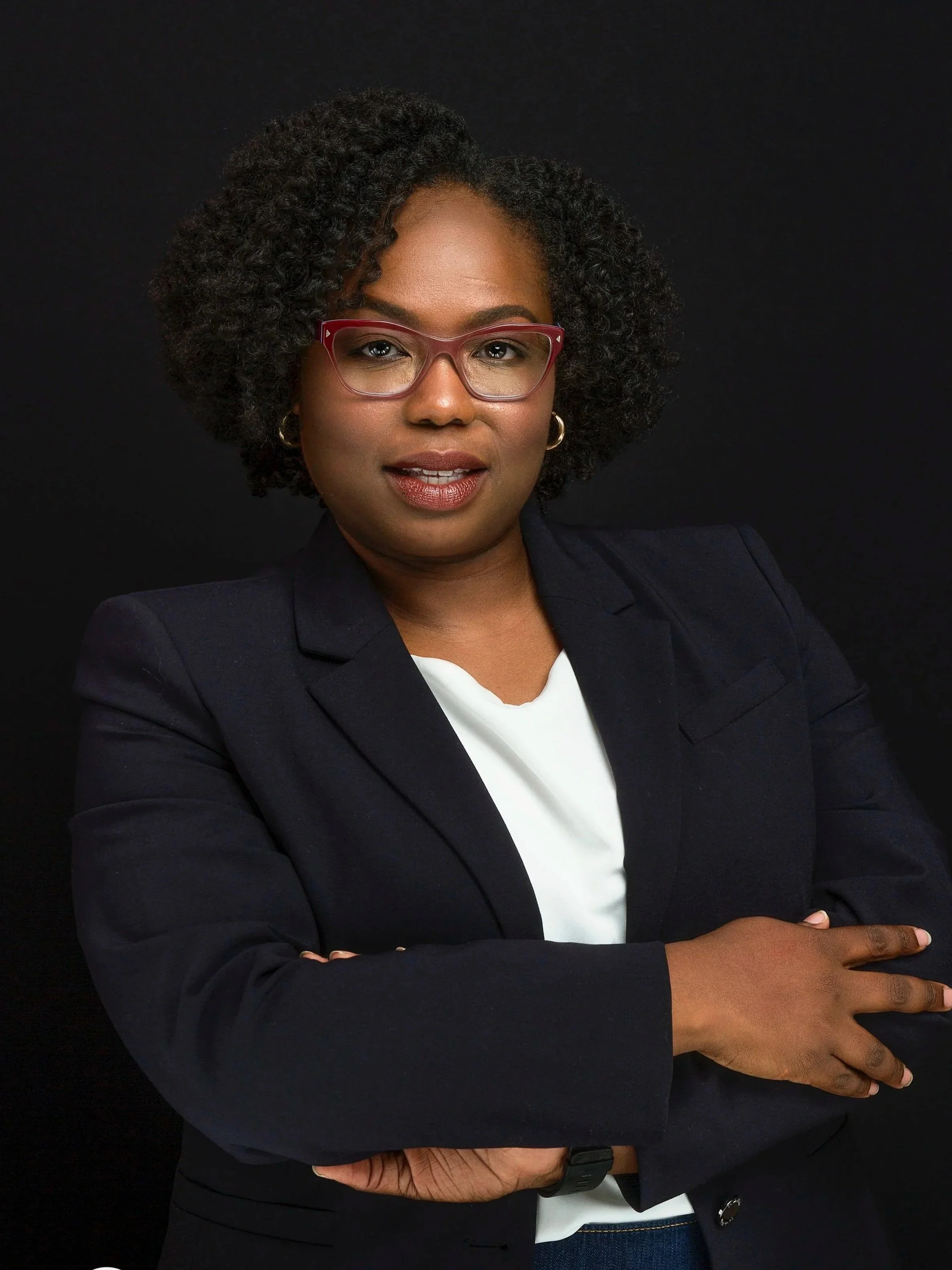 A woman with dark skin, curly hair, pink glasses, wearing a dark blazer and white shirt, standing against a black background.
