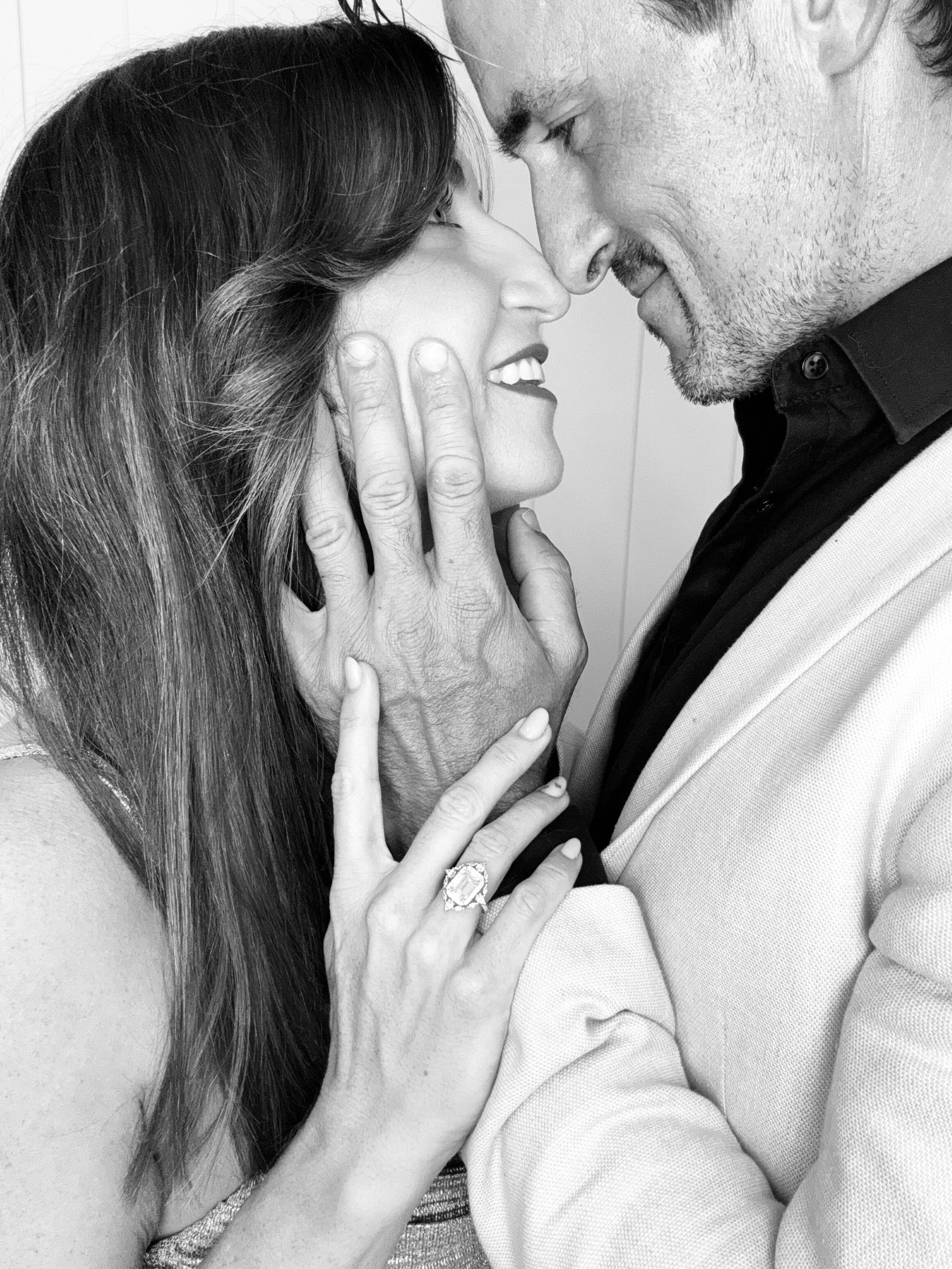 A black and white photo of a happy couple with their foreheads and noses touching, smiling warmly at each other. The woman has long hair and is wearing a ring on her finger. The man has short hair and is wearing a light jacket over a dark shirt.