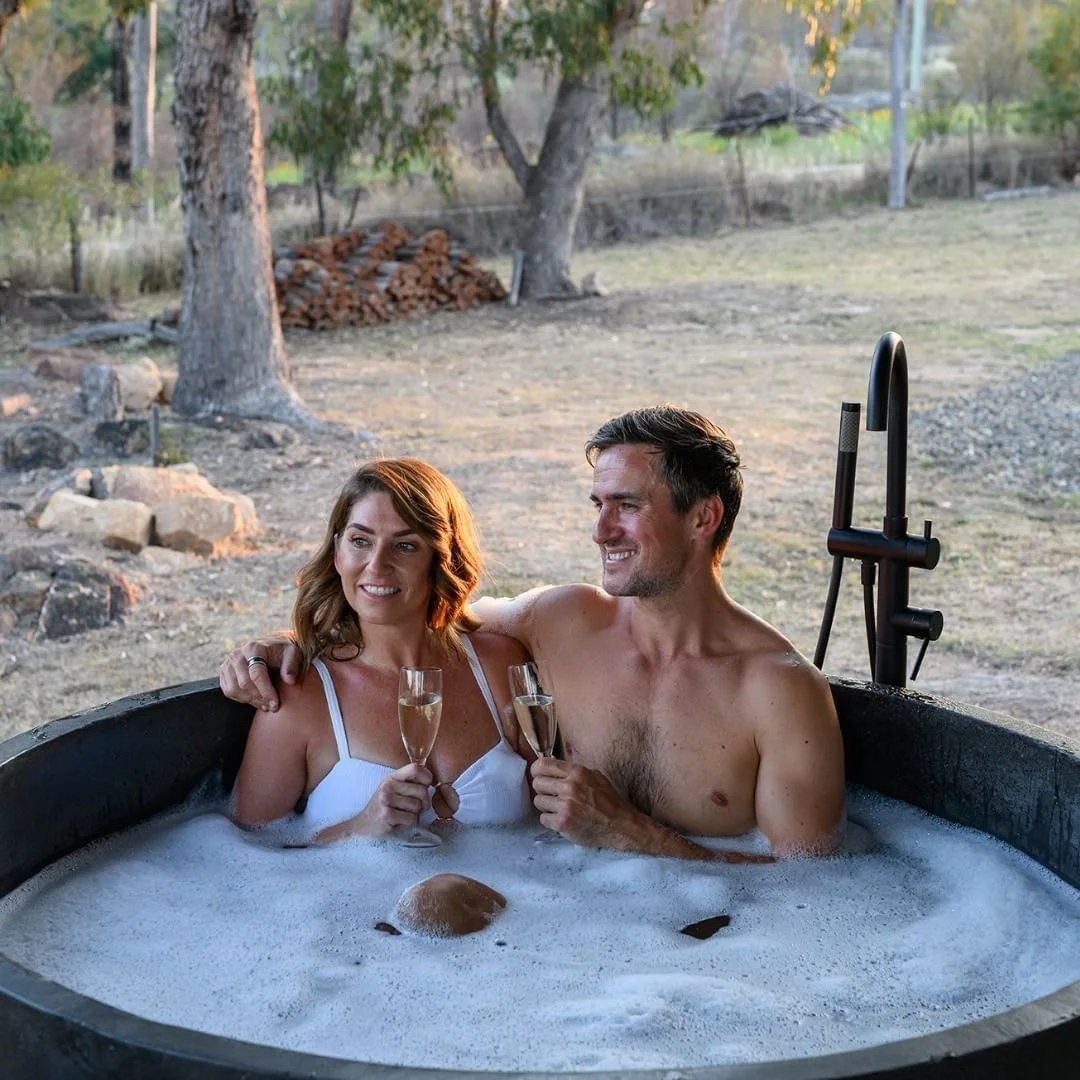 A couple enjoying a bubble bath together outdoors, holding champagne glasses and smiling.