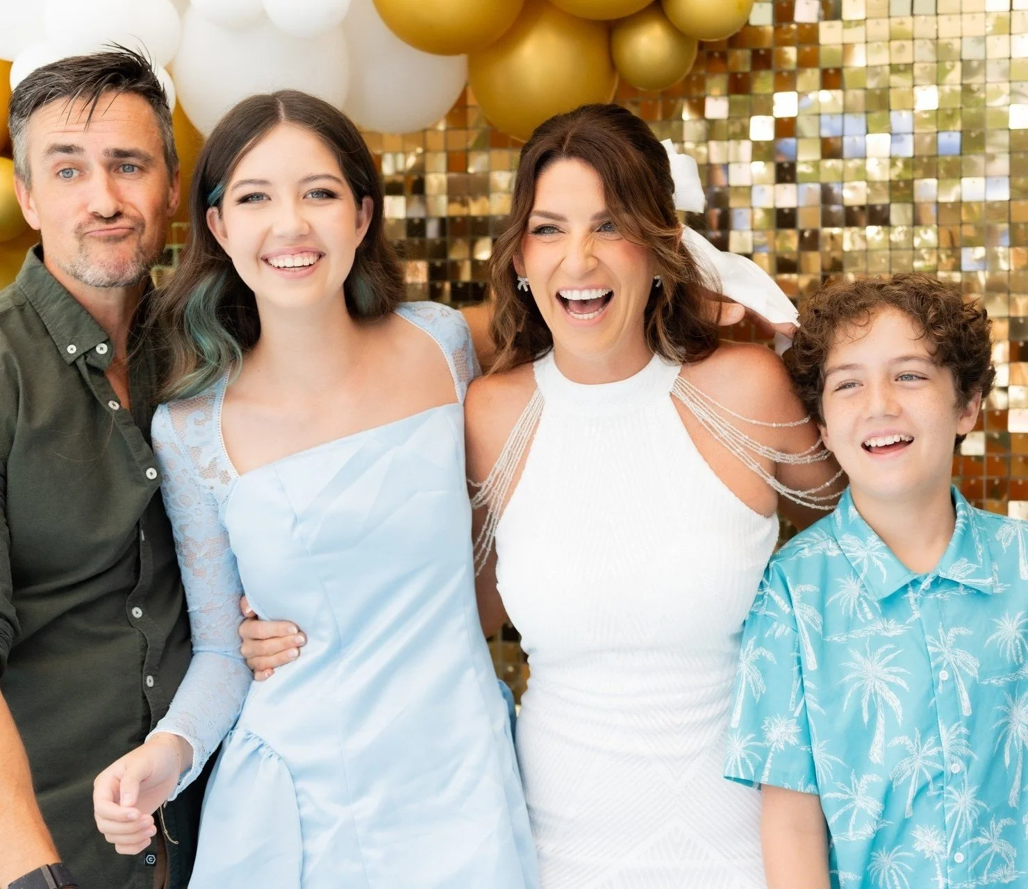 A group of five smiling people, two women, one man, and two children, at a celebration with balloons and a golden tiled backdrop.