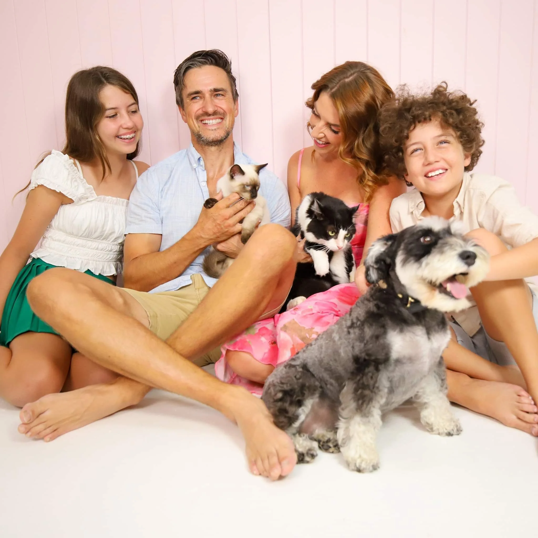 Family sitting on the floor with their pets, including cats, a dog, and a puppy, in a cozy setting.