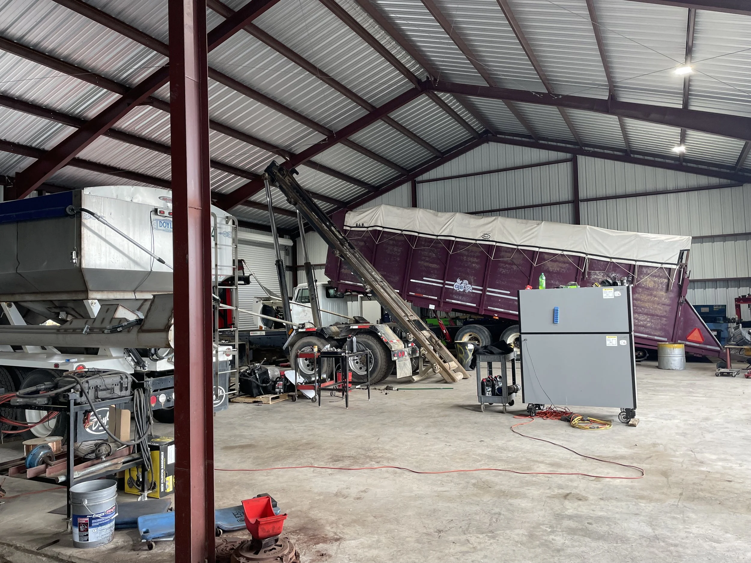 Indoor garage or workshop with large maroon and white trailer, various tools, equipment, and parts scattered around, and a high metal ceiling with beams.