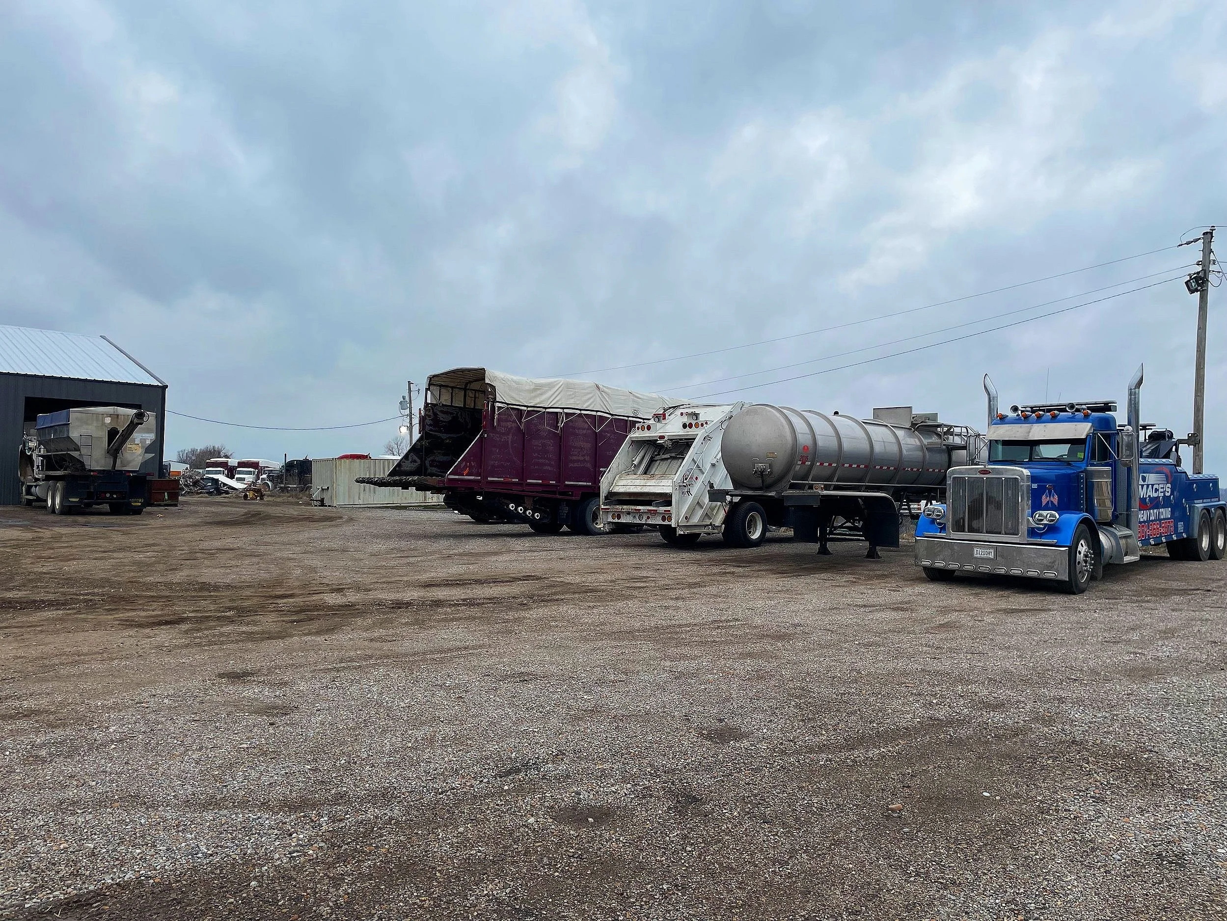 A blue semi-truck parked in a lot with gravel ground, alongside tankers, trailers, and cloudy sky.