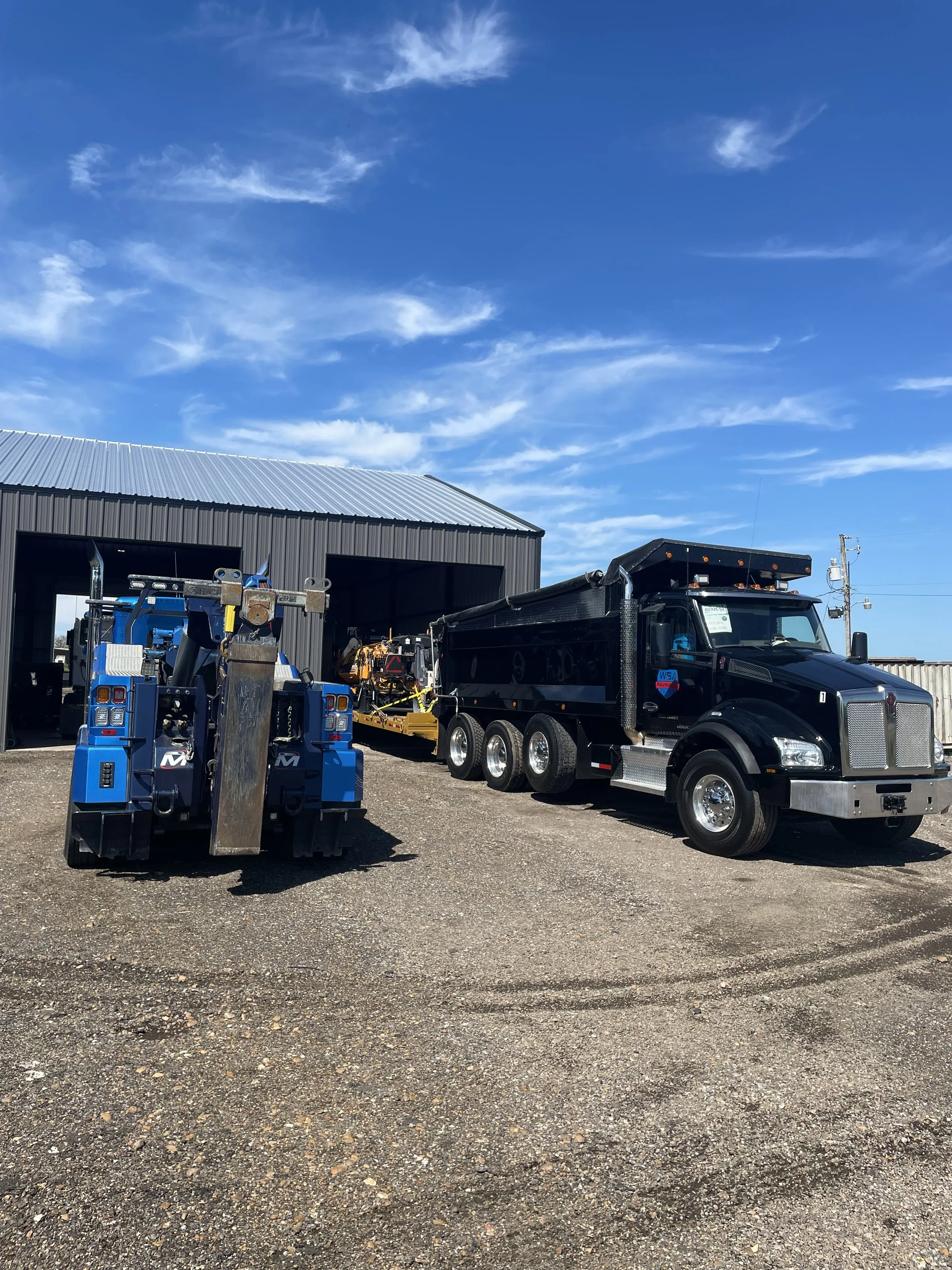 A large black semi-truck parked outside a black metal building with a blue sky and clouds overhead. The truck is attached to a yellow piece of equipment and a large blue vehicle is visible in front.