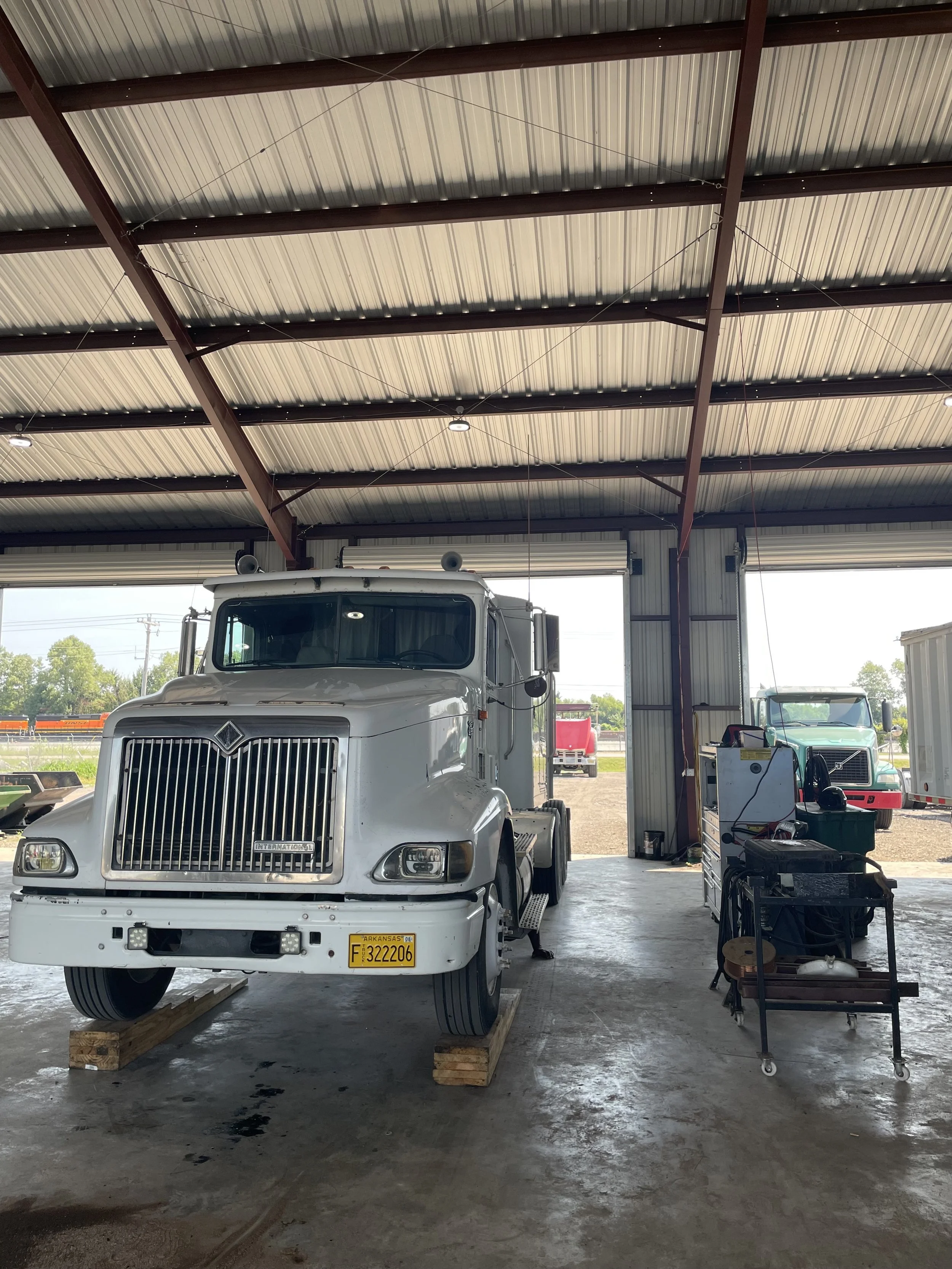 Inside a shop with a white semi-truck parked on wooden blocks, and a smaller truck and workbench with tools in the background.