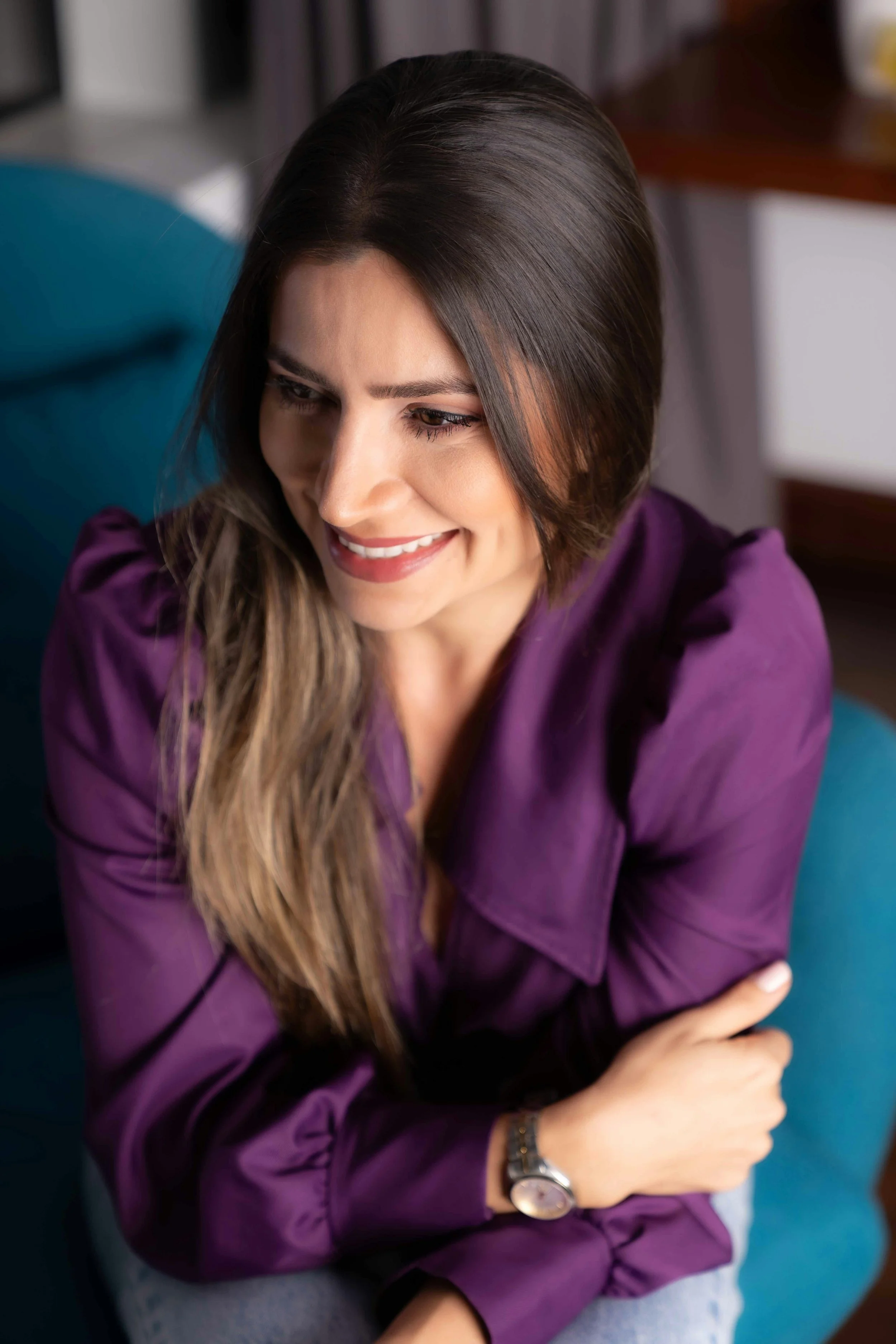A woman with long brown hair wearing a purple blouse and a watch, sitting on a teal chair, smiling and looking down.