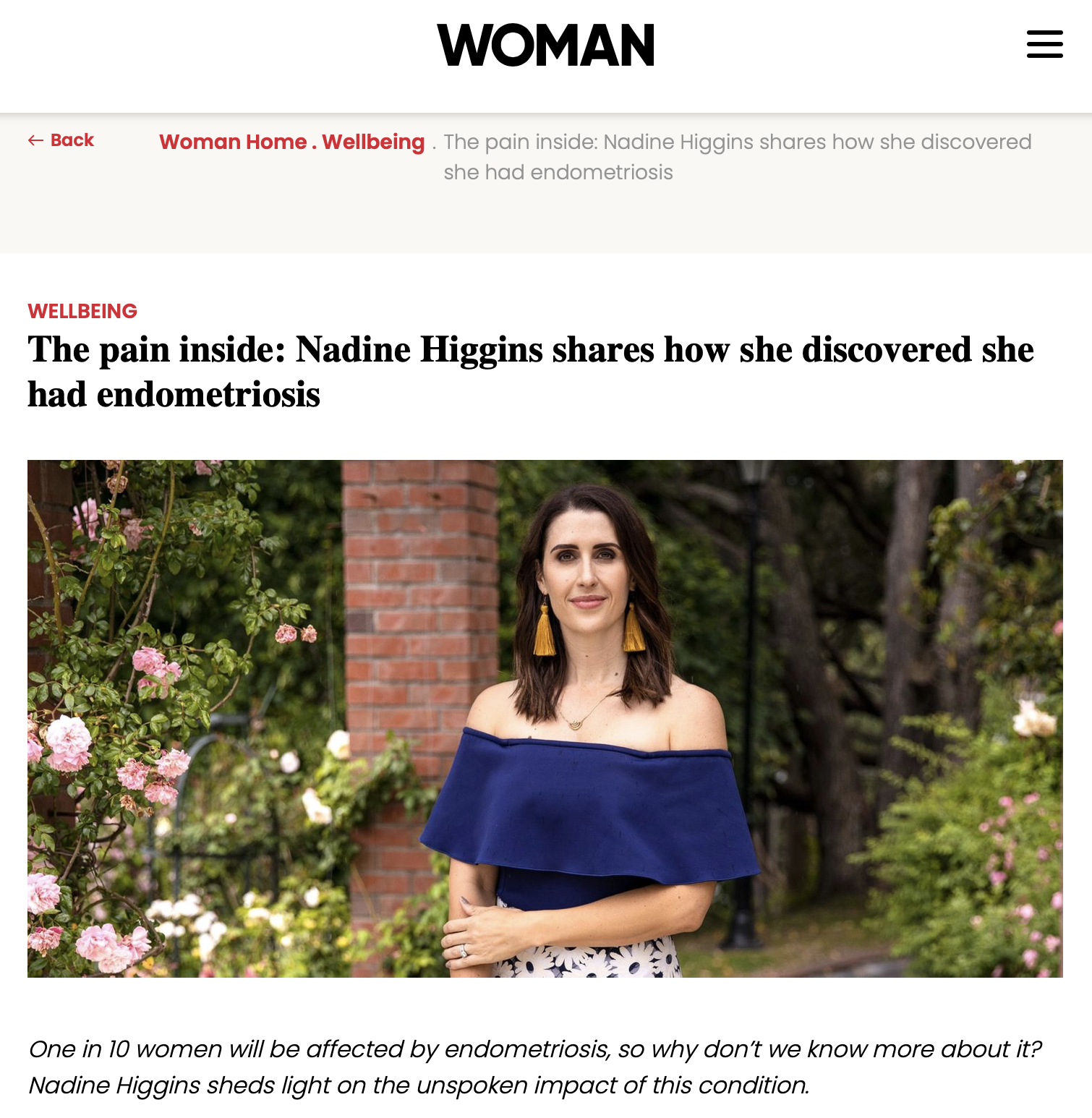 A woman with shoulder-length brown hair, wearing a royal blue off-the-shoulder top and yellow tassel earrings, standing outdoors in a garden with pink roses and a red brick pillar.