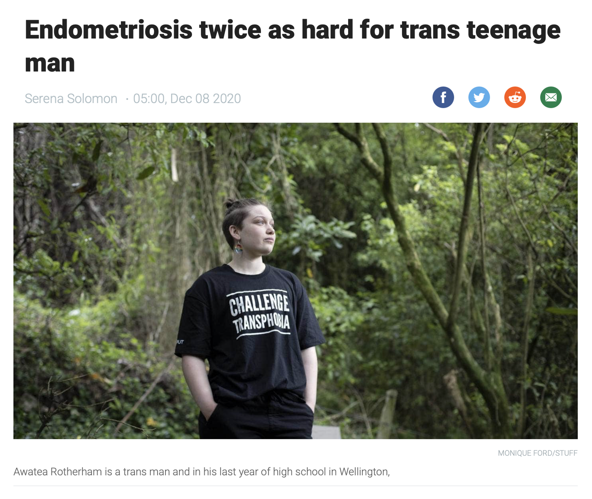 Transgender teenager standing outdoors in a green forested area, wearing a black T-shirt with white text that says 'Challenge Transphobia', with hands in pockets, looking off to the side.