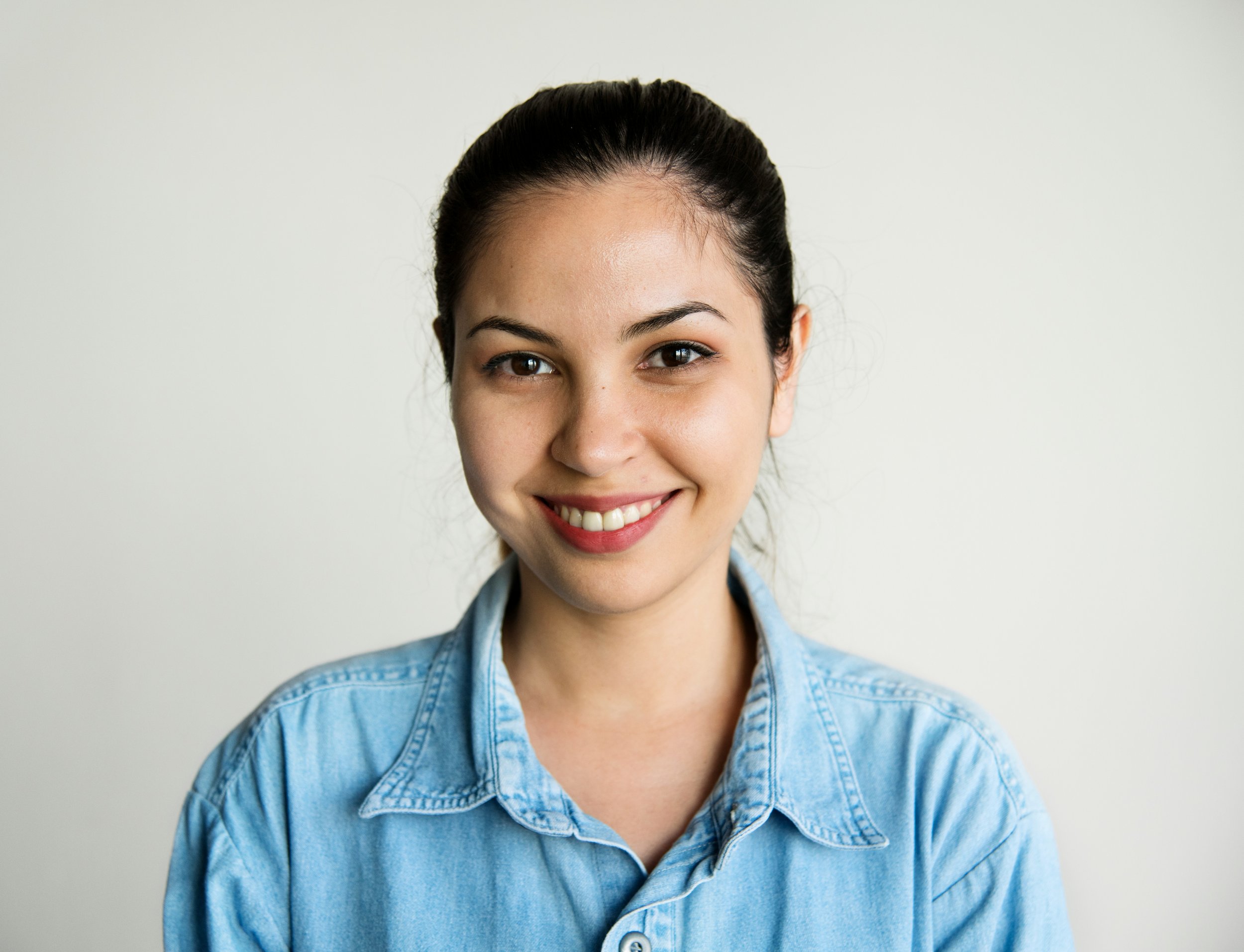 A woman with dark hair tied back, wearing a blue denim shirt, smiling and looking directly at the camera against a plain, light-colored background.