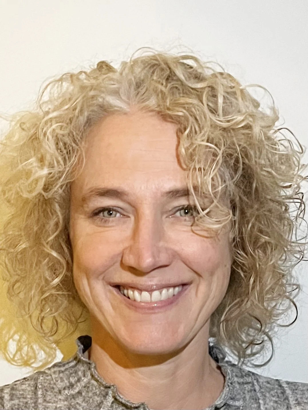 Close-up of a woman with curly blonde hair, smiling, against a light-colored background.