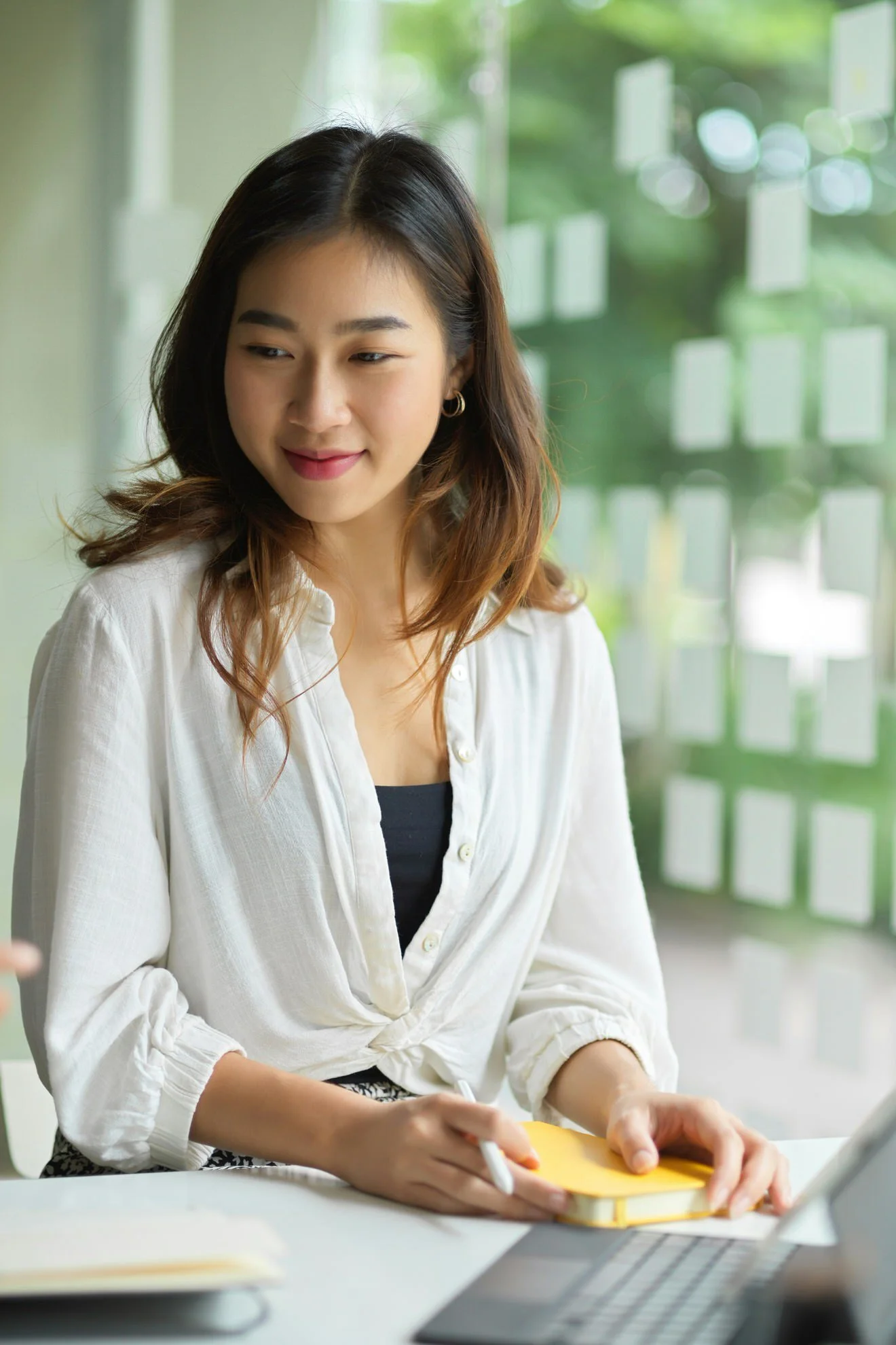 A young woman sitting at a desk with a laptop, taking notes on a yellow notepad.