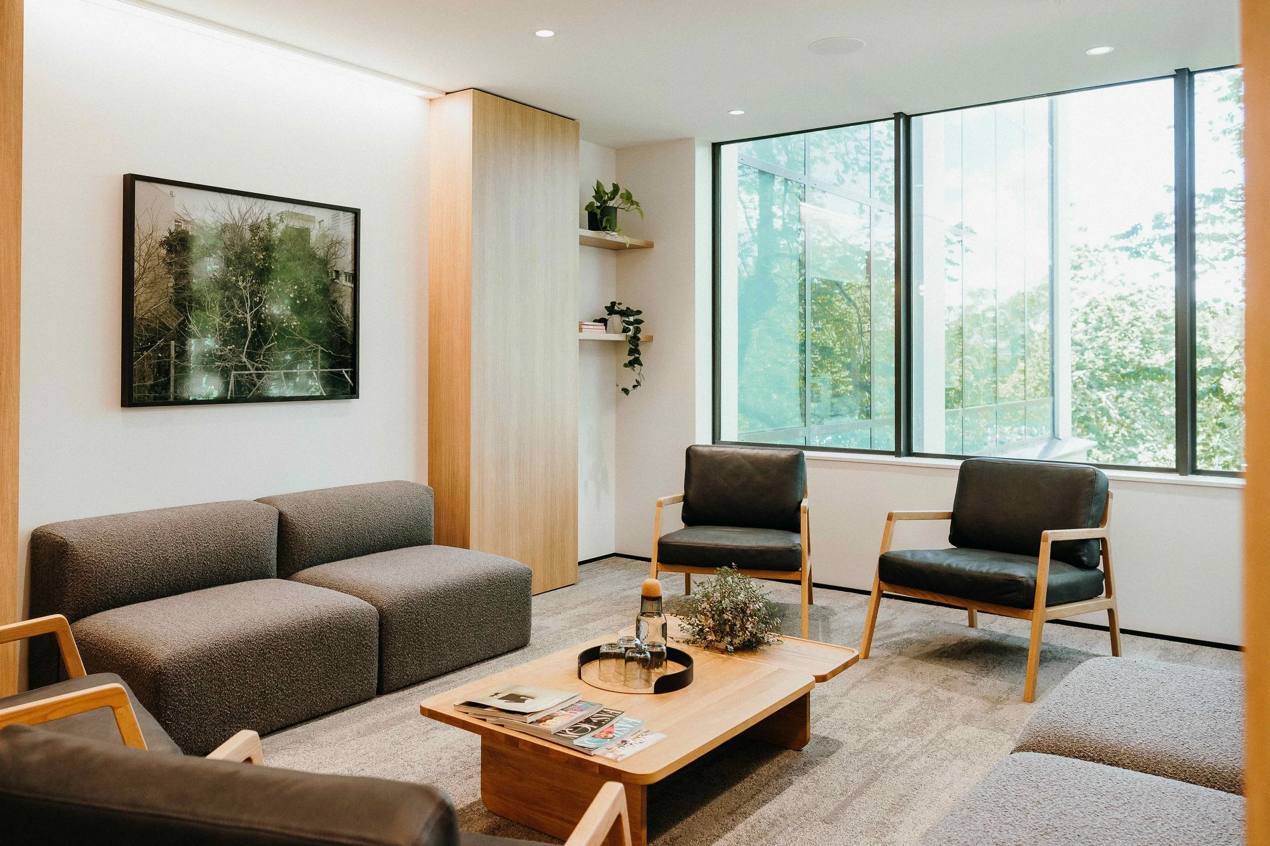 Modern living room with gray sofa, two black armchairs, a wooden coffee table with magazines and a plant, large windows with greenery outside, and shelves with plants and books.