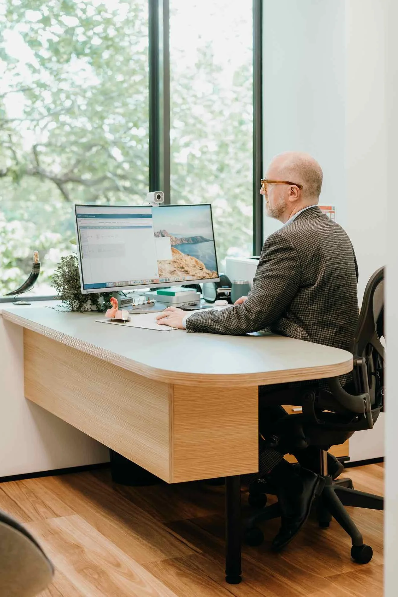 A man with glasses and a beard working at a wooden desk in an office, using a dual monitor setup with a webcam on top, in front of large windows with greenery outside.