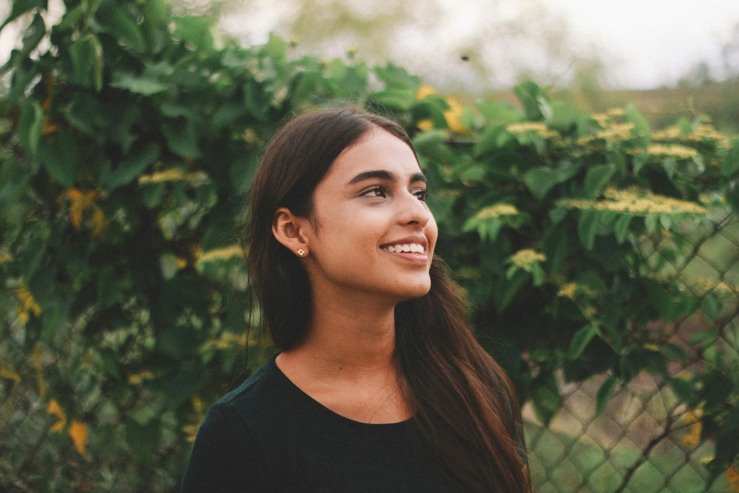 A young woman with long dark hair, smiling and looking to the side, standing outdoors in front of green foliage and a chain-link fence.