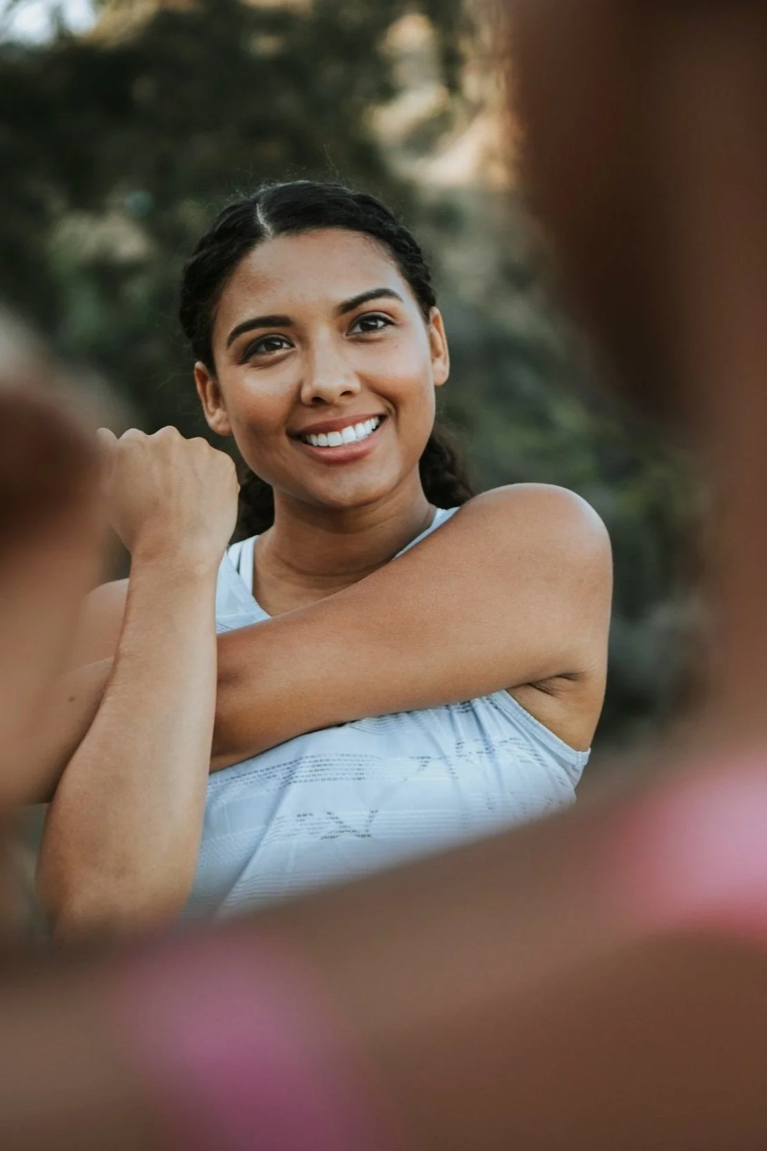 A young woman smiling outdoors, with her arm raised and her other arm crossed over her chest, surrounded by out-of-focus individuals.