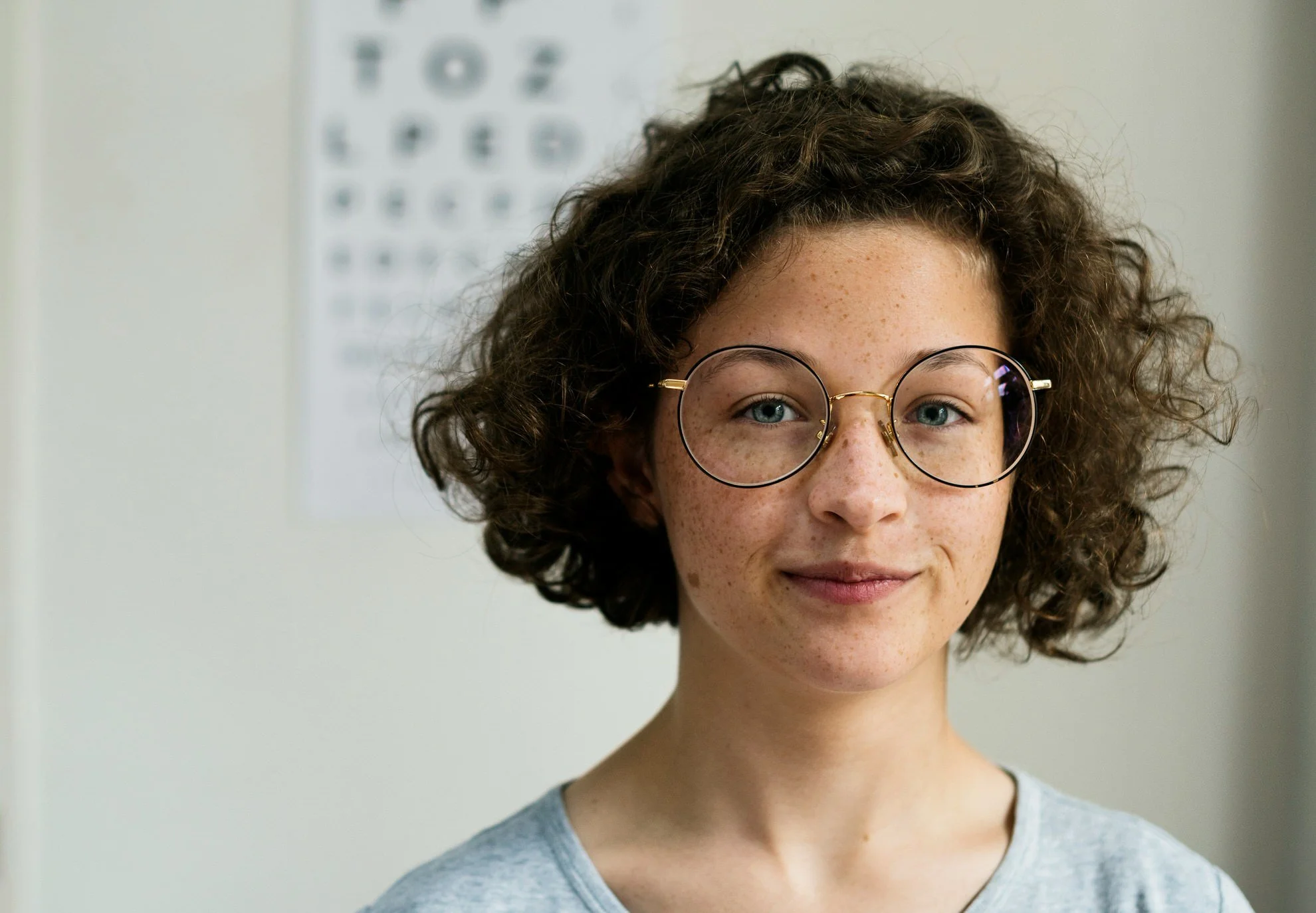 A young girl with curly brown hair, wearing round glasses and a gray shirt, standing in front of an eye chart on the wall.