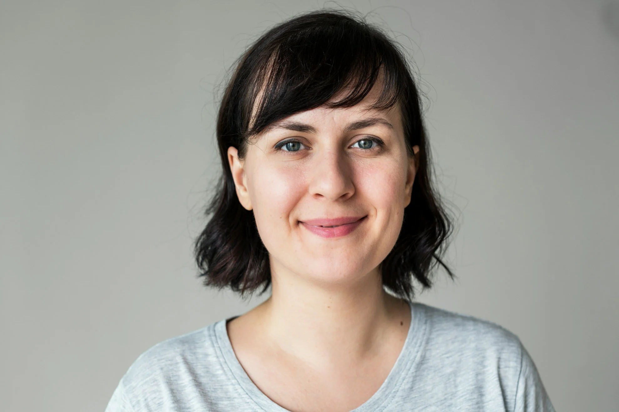 A young woman with shoulder-length dark hair and blue eyes smiling at the camera, wearing a gray t-shirt against a plain gray background.