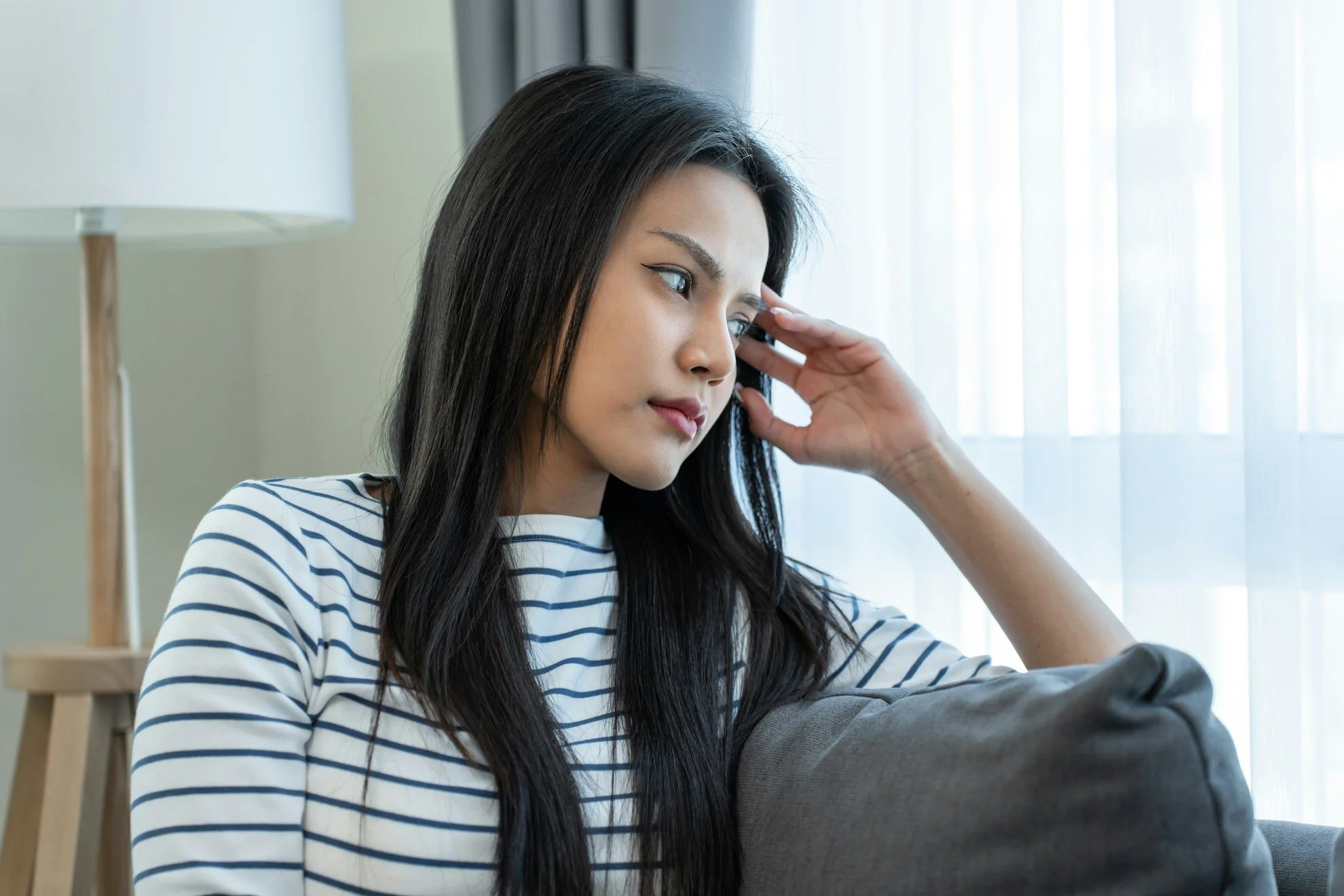 A young woman with long dark hair wearing a striped shirt sitting on a couch near a window, appearing thoughtful or pensive.