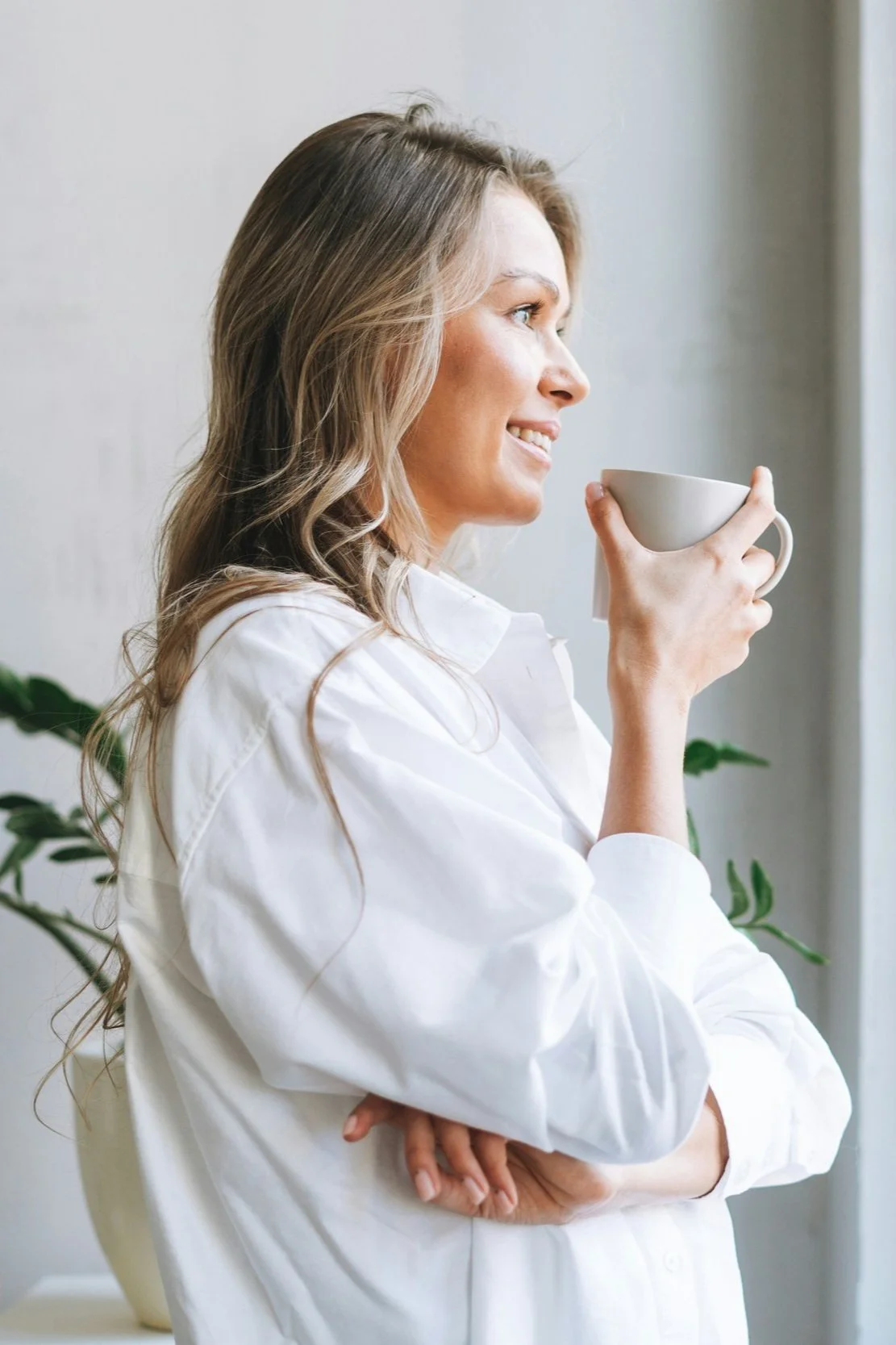 A woman with long hair and a white shirt holding a white mug near her face, smiling as she looks out of a window.