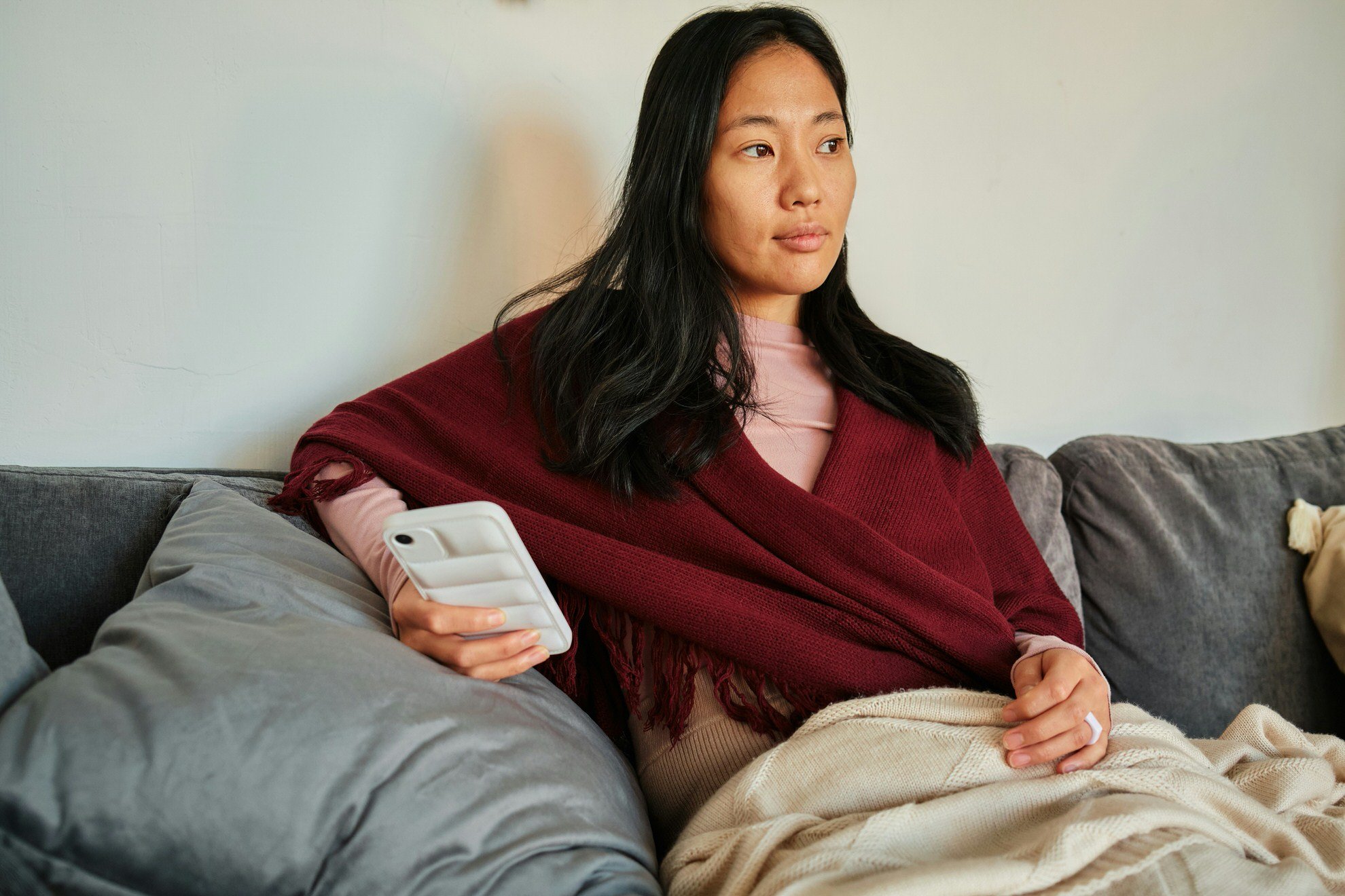 Woman sitting on a gray couch, holding a smartphone in her right hand, wrapped in a red shawl, looking thoughtfully into the distance.