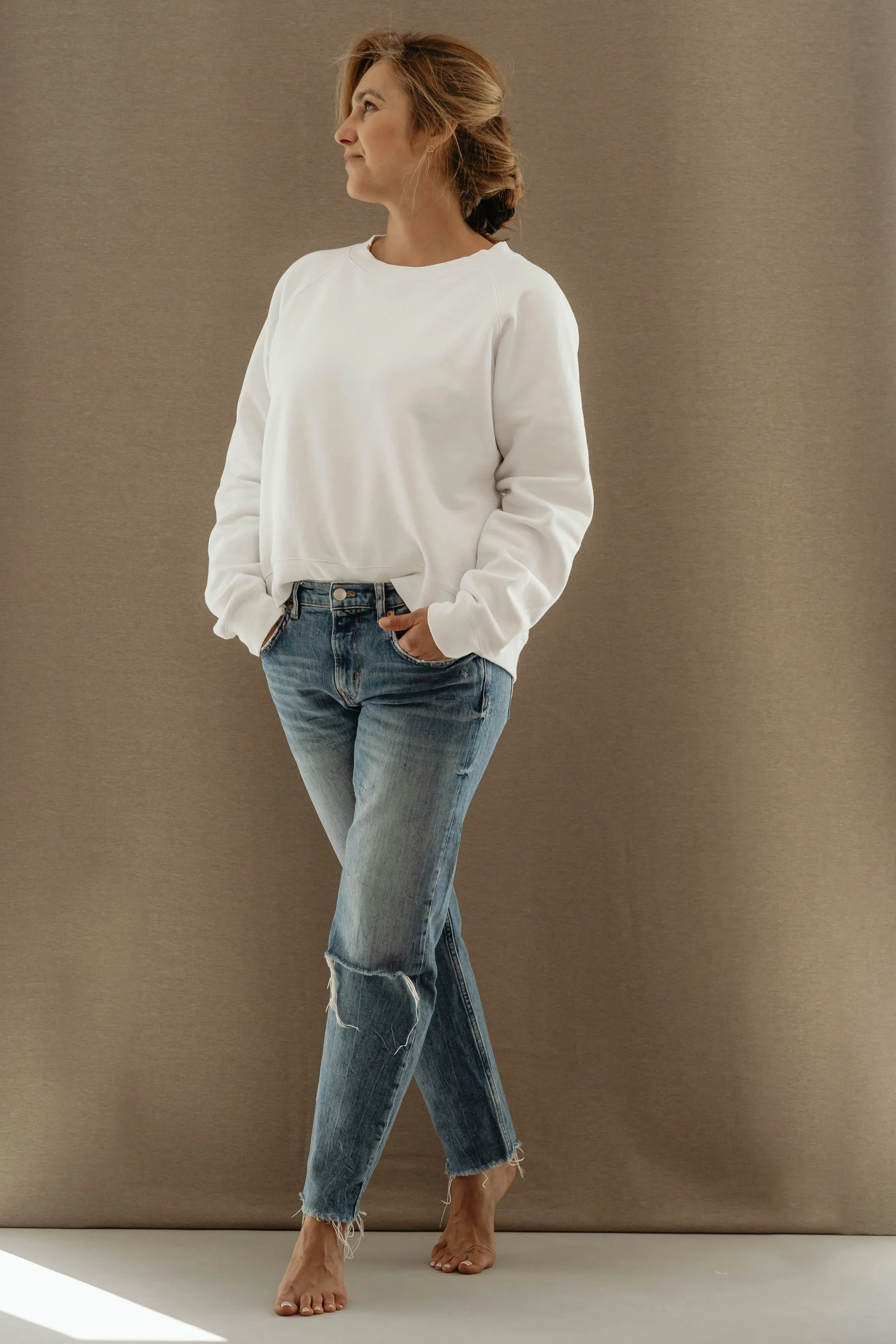 Woman with light brown hair in a ponytail wearing a white sweatshirt and distressed blue jeans standing barefoot against a beige background.