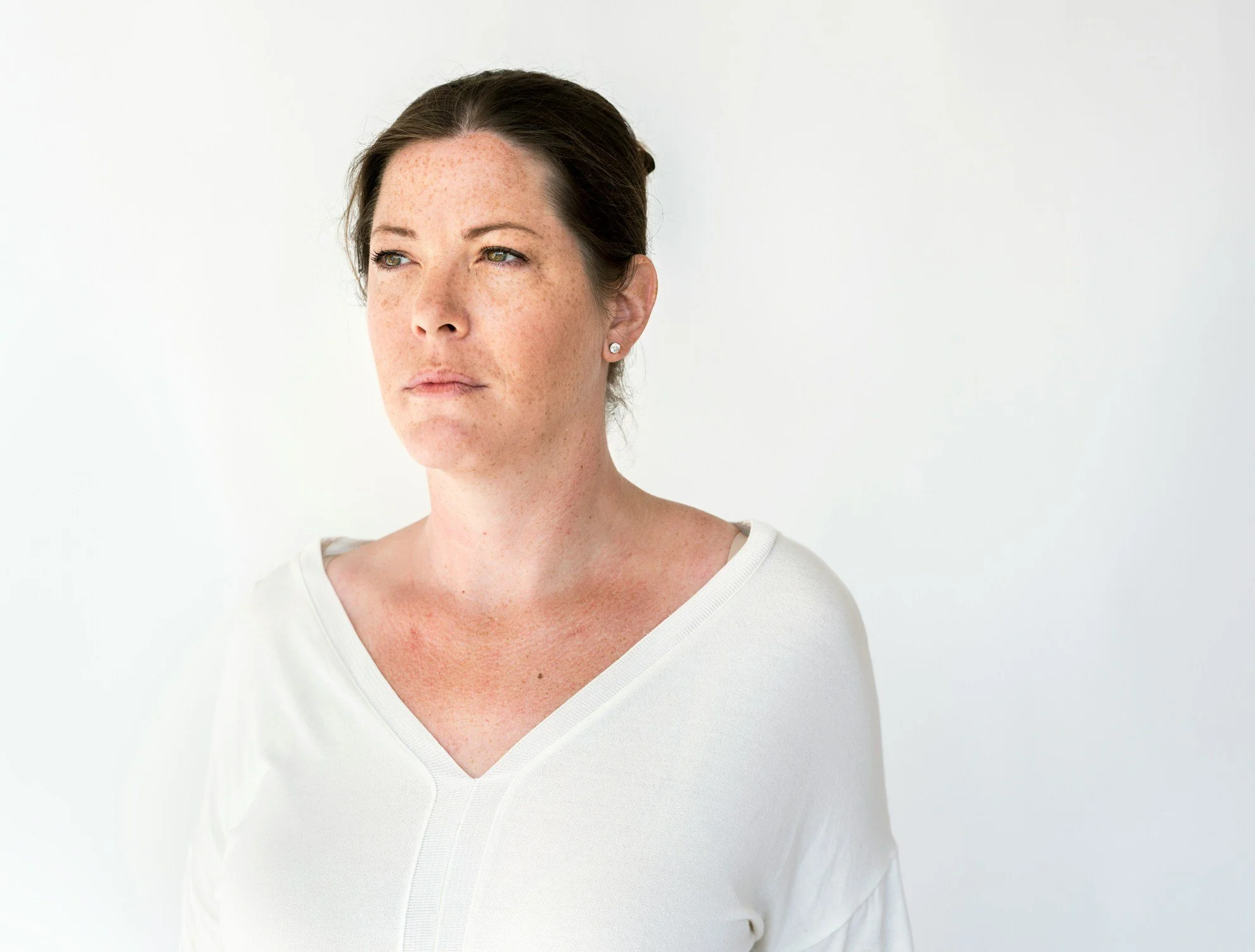 Close-up of a woman with dark hair, light skin with freckles, wearing a white top against a plain white background, looking to the side.