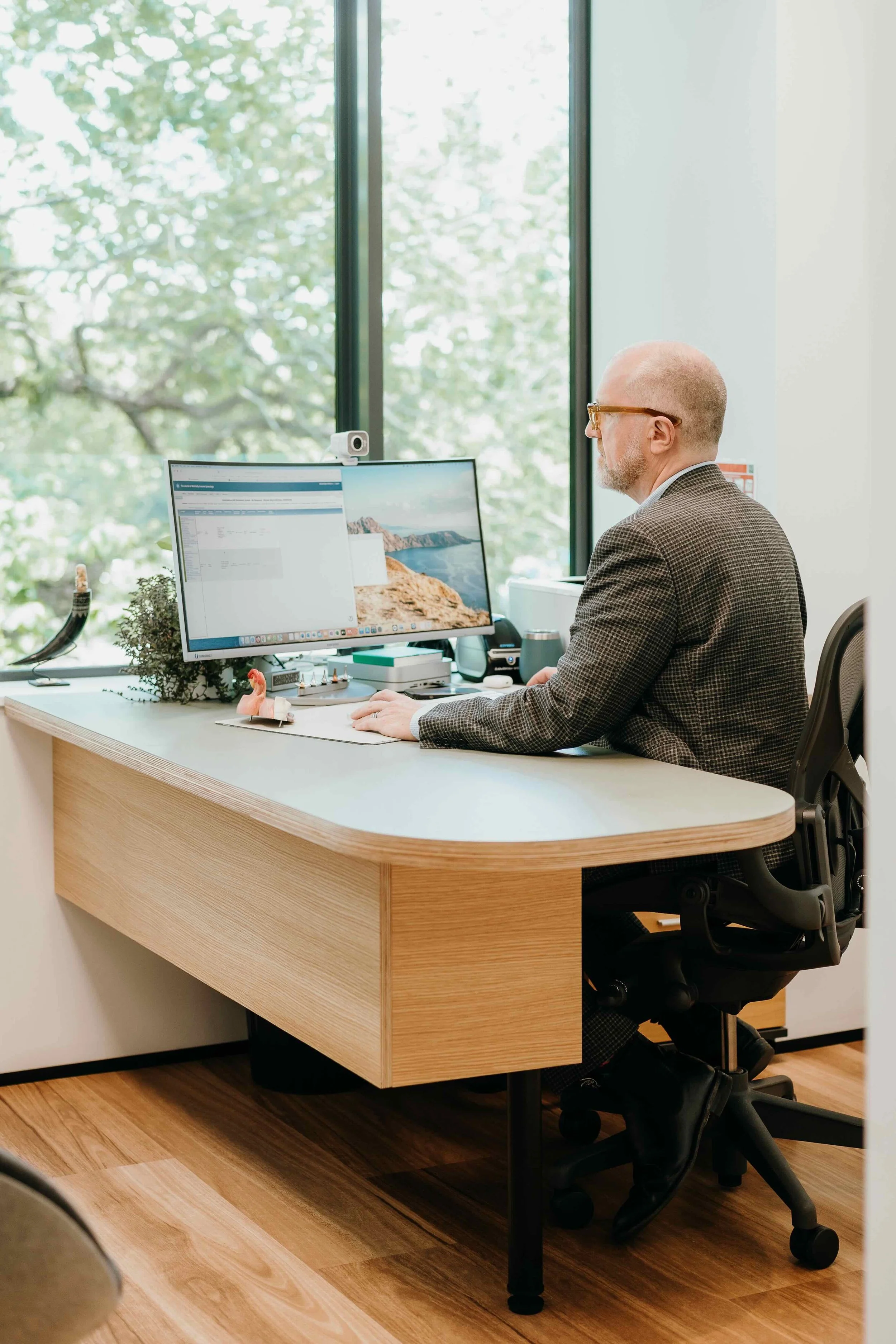 Man with glasses in a suit working at a desk in an office with a large window showing green trees outside