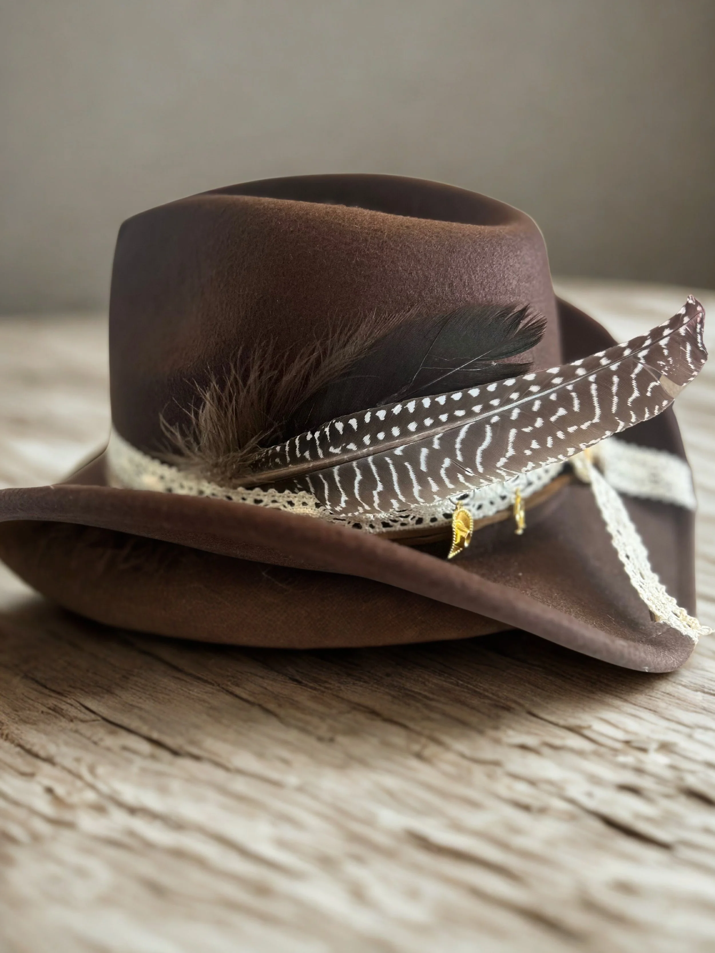 Brown felt cowboy hat decorated with a large black feather, a spotted feather, a lace band, and small gold charms, placed on a wooden surface.