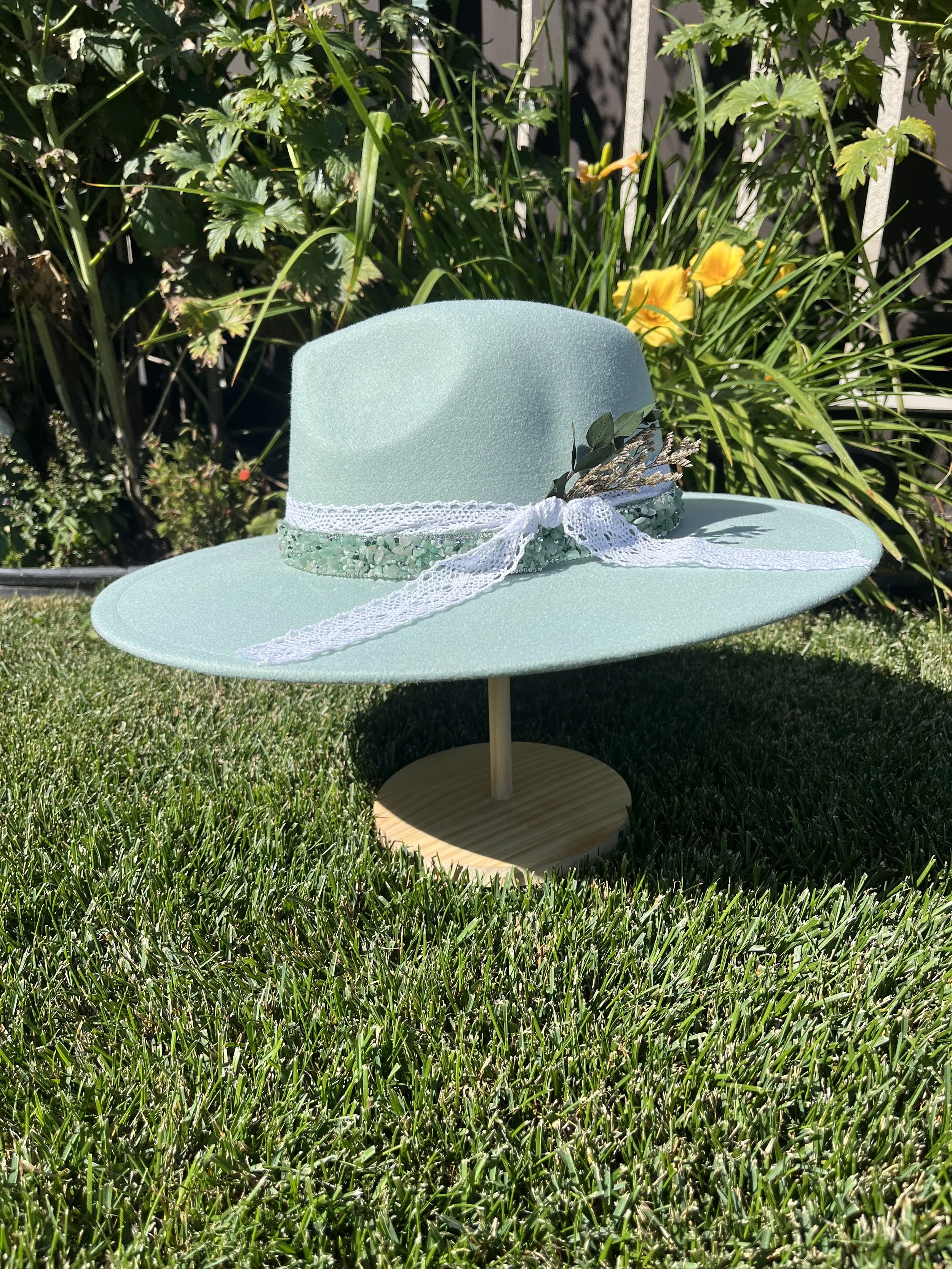 A light green wide-brimmed hat with lace, ribbon, and greenery decoration, placed on a wooden stand on the grass in a garden with green plants and yellow flowers in the background.