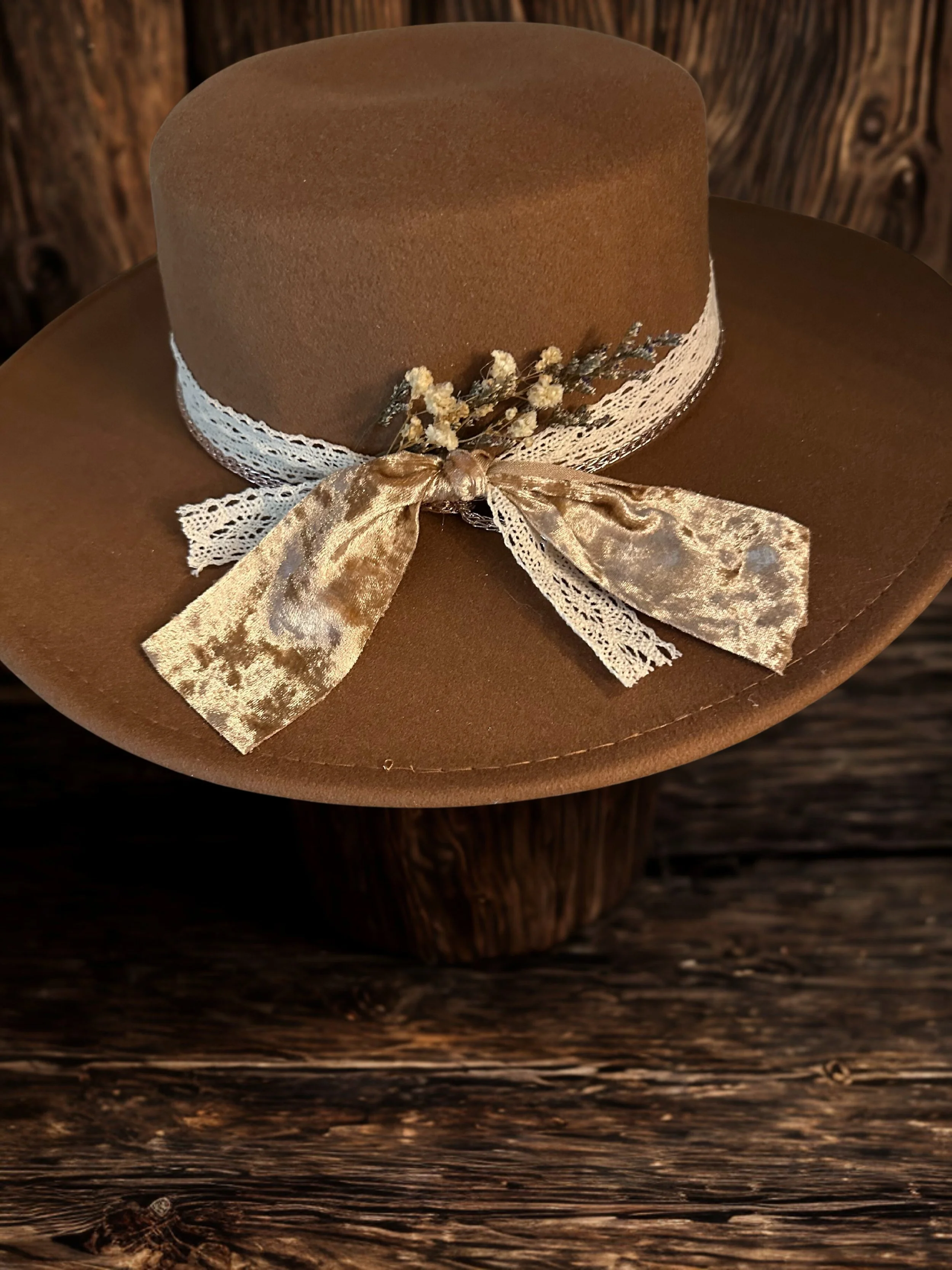 A brown cowboy hat decorated with a lace ribbon, a small flower arrangement, and a satin bow, sitting on a rustic wooden surface.