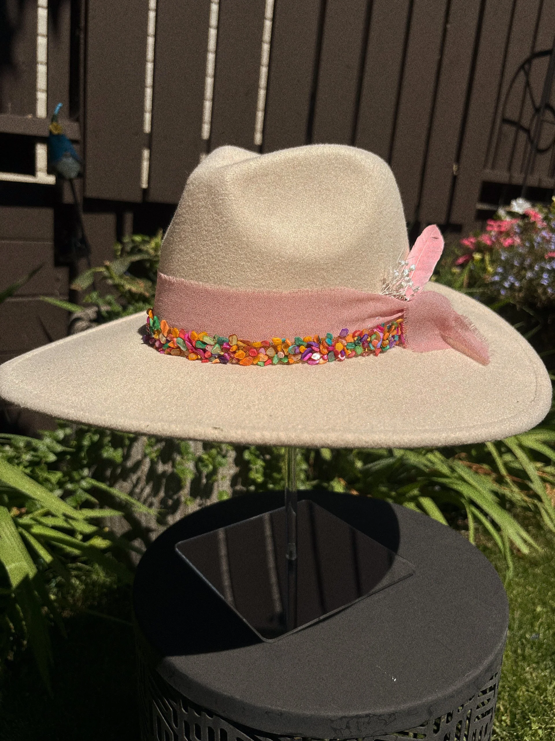 A beige wide-brimmed hat with a pink band and multicolored embellishments, displayed outside on a stand against a background of plants and flowers.