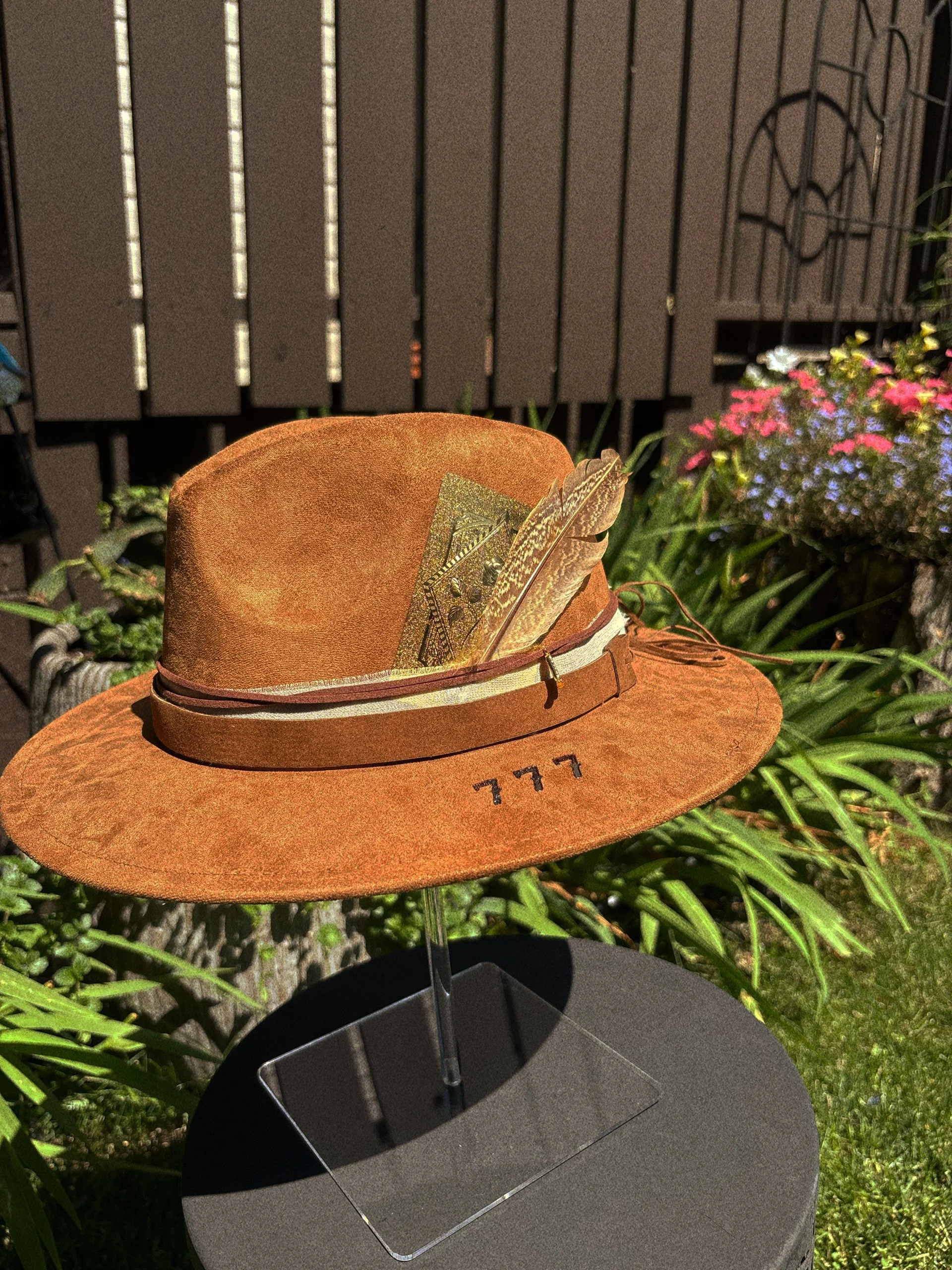 A tan cowboy hat with a white band and three black tally marks embroidered on the brim, decorated with two feathers, displayed outdoors among green plants and colorful flowers with a wooden fence in the background.