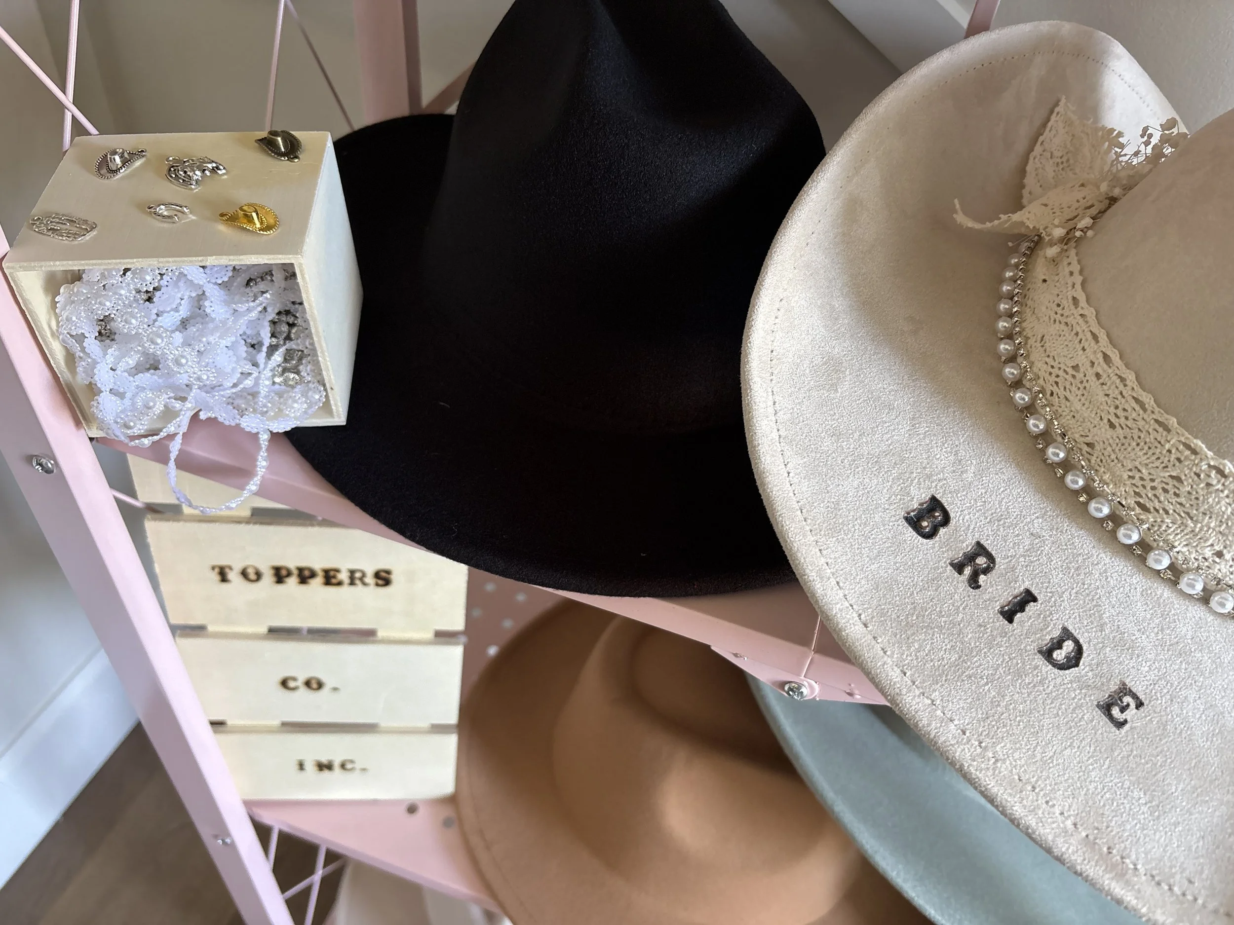 A display of four cowboy hats on a pastel pink rack, with a small cream-colored box containing jewelry and pins on top. The hats are labeled 'BIRDIE' and the rack has a sign saying 'TOPPERS CO.'