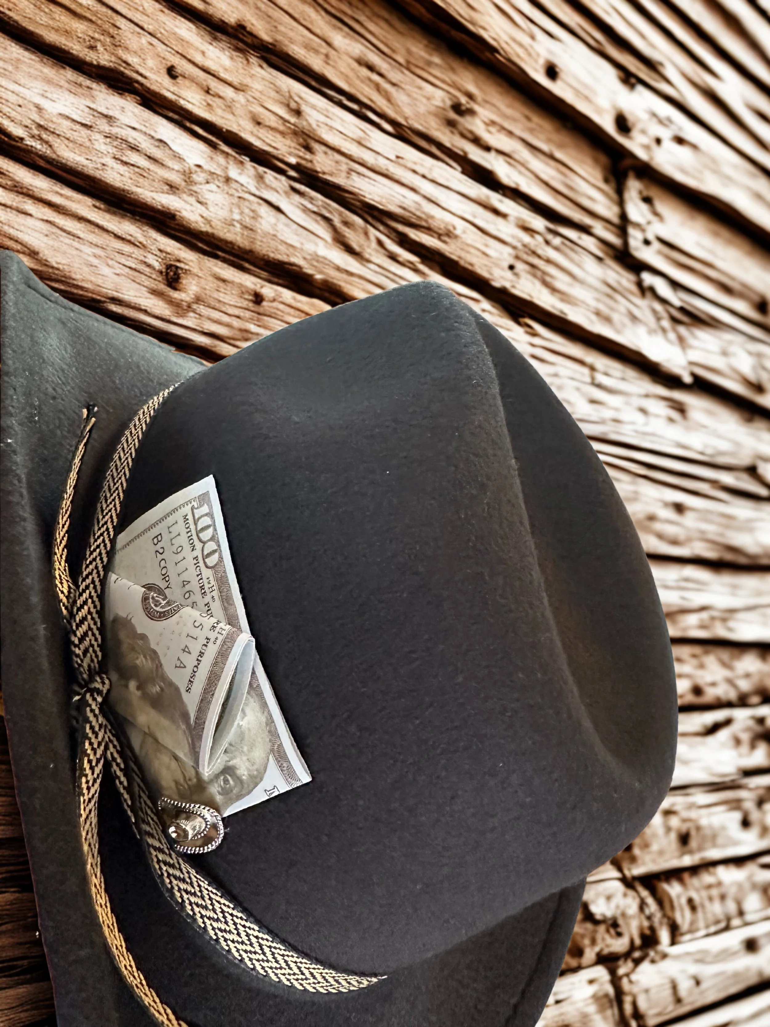 A black felt hat with a dollar bill inside its folded brim and a black and white braided hatband, resting on a wooden surface with weathered, textured planks.