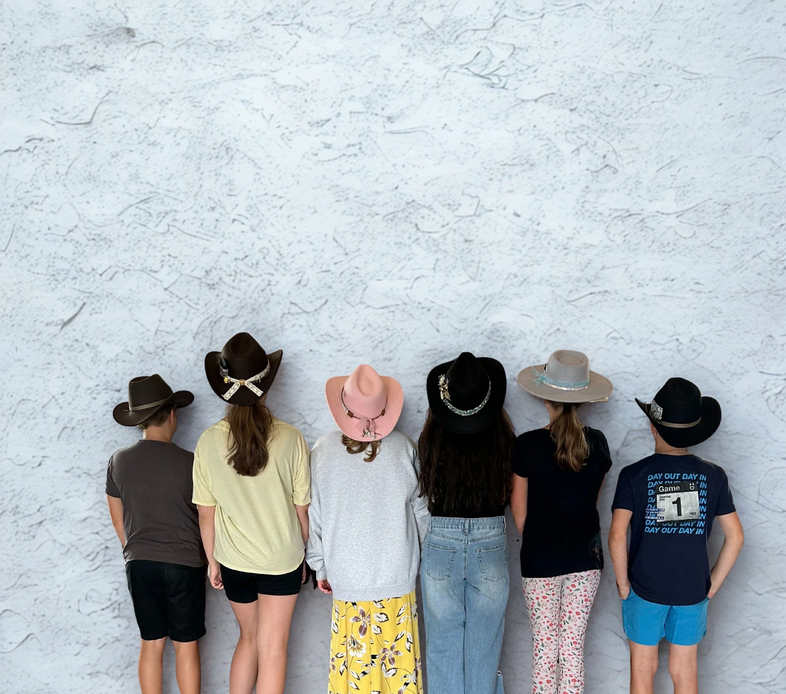 Six children wearing hats, standing side by side with backs facing the camera, against a textured white wall.